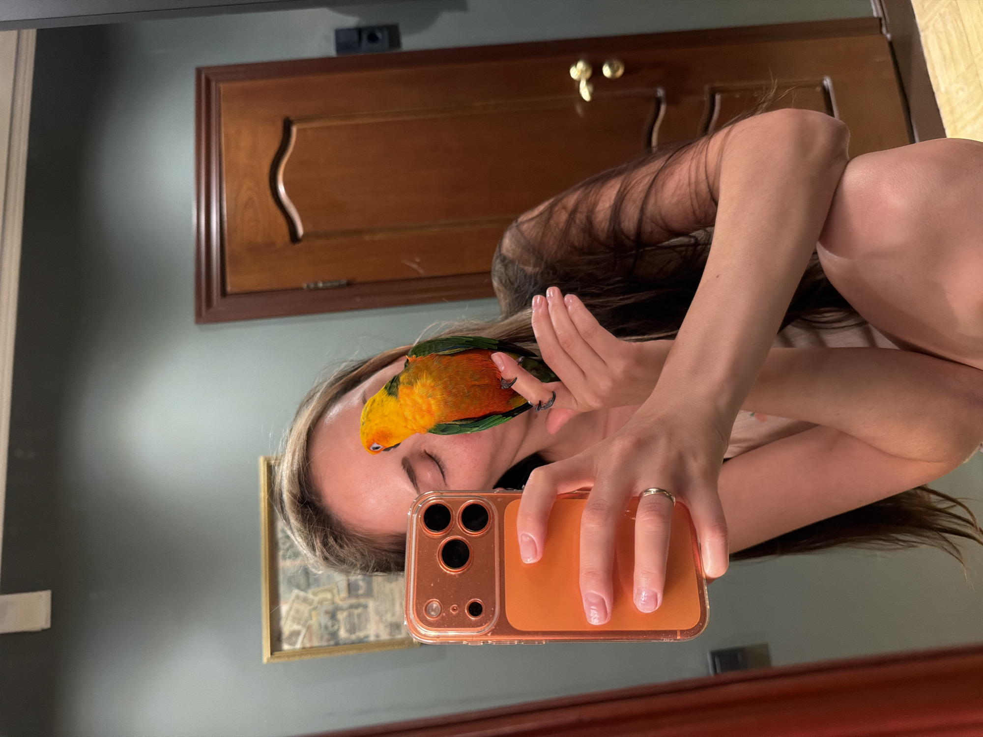 A woman is taking a selfie in a mirror with a parrot perched on her head. She is holding an orange-cased iPhone. Her left hand is covering her face. She has light skin and long brown hair. The parrot is orange and yellow with green wings. Behind her, the wall is a gray-green color, with a wooden door with gold hardware, and a framed picture on the wall. I cannot determine a location in Khimki, Russia from the image.