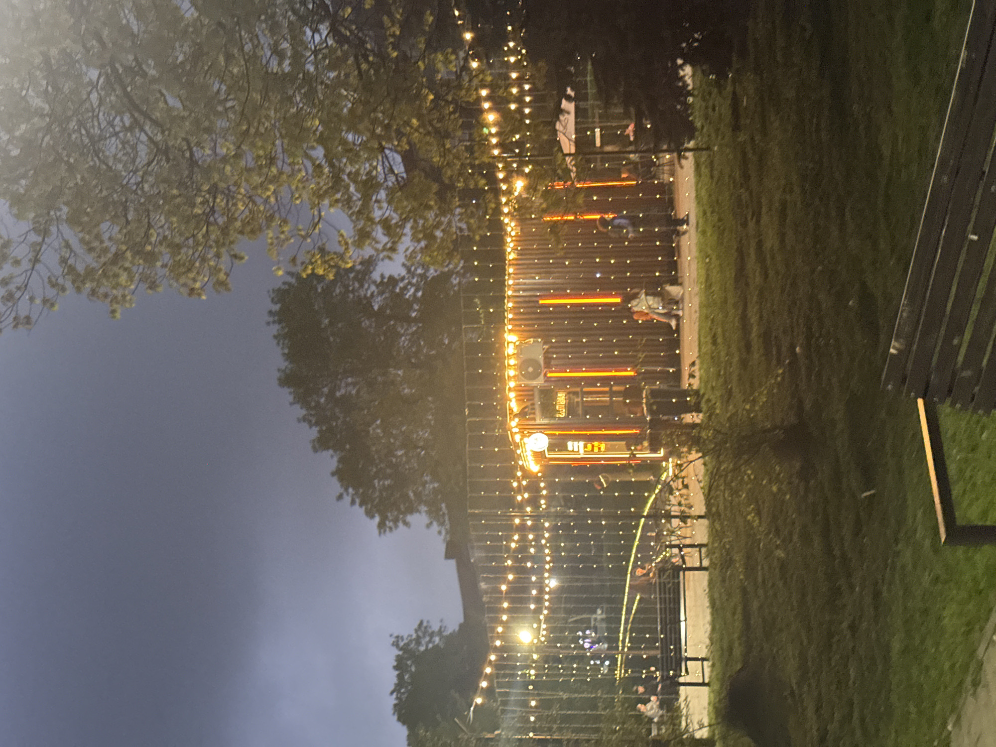 A nighttime scene depicts an outdoor area. A building or structure illuminated with orange and white lights is partially visible, adorned with multiple strings of small lights. Trees are present on the left and in the background, silhouetted against a dark sky. On the right, a section of a wooden bench is visible, with grass and possibly a pathway extending into the background. People are seated and standing in front of the illuminated building. This is Komgaron, Russia.