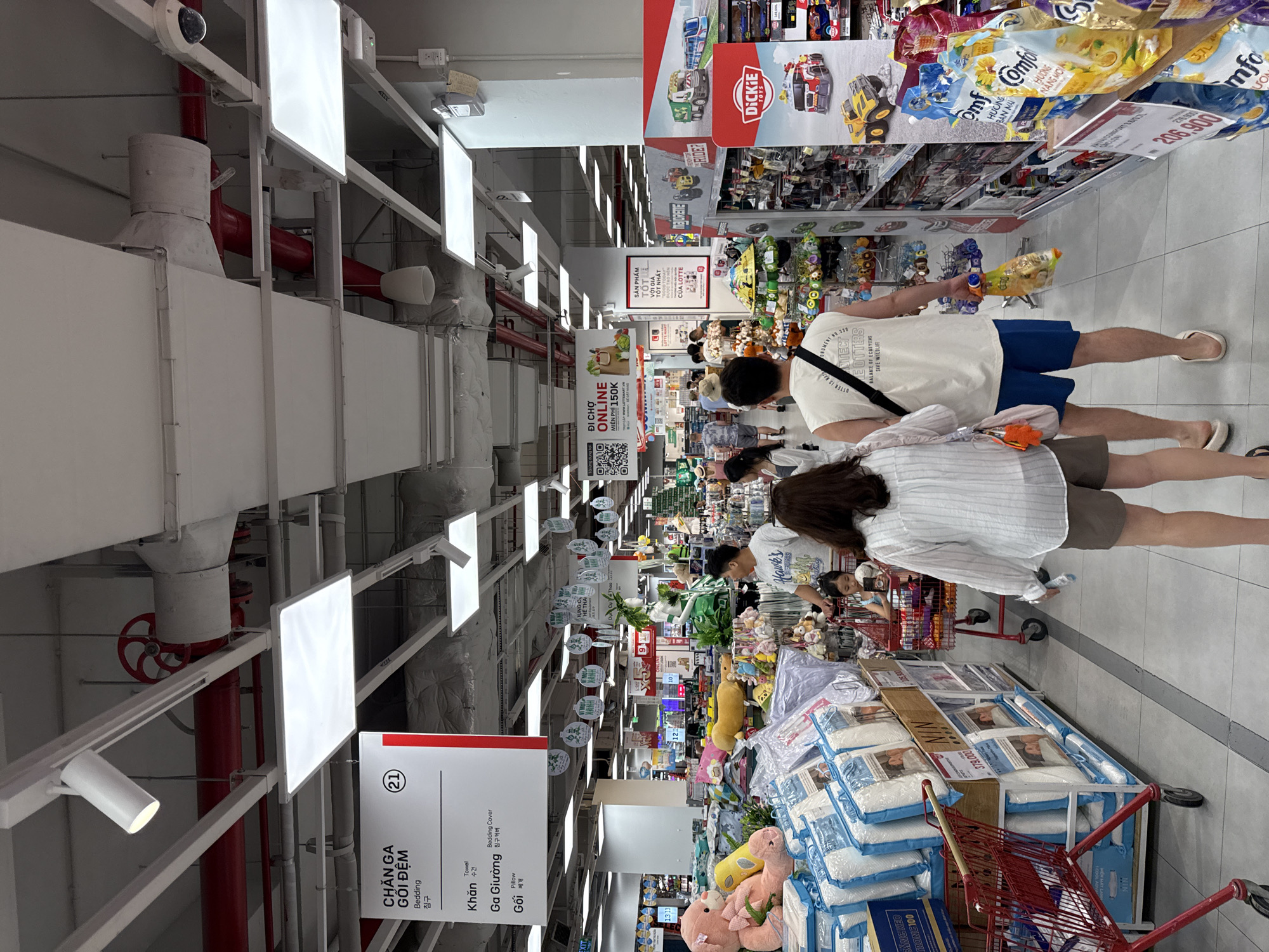 The image captures an interior view of a large retail store, likely a hypermarket, in Nha Trang, Vietnam. The setting is brightly lit with a combination of overhead fluorescent and spotlighting. The environment appears to be a section dedicated to home goods and possibly toys, with aisles of merchandise visible.
In the foreground, two individuals are walking away from the camera, down an aisle. The person on the left is wearing a white, vertically striped shirt and brown shorts, carrying a purse. The person on the right is wearing a white sleeveless t-shirt with text on it and blue shorts, carrying a bag of items. Further down the aisle, other shoppers and carts are visible.
Merchandise is displayed on shelves and on the floor. In the left portion of the image, large stacks of packaged bedding items, predominantly in blue and white, are prominent. A red shopping cart is parked to the left of these displays. In the middle and right sections, shelves are stocked with various products, including what appear to be toys and household items. A prominent display on the right features toy trucks from the "Dickie Toys" brand.
Signage in Vietnamese is visible throughout the store, including a red and white sign at the top left labeled "CHĂN GA GỐI ĐỆM" (Bedding) with further product categories listed. Another sign further down the aisle promotes "ONLINE" shopping with a price of "MIỄN PHÍ 150K" (Free for 150,000 VND). The overall atmosphere suggests a busy shopping day.