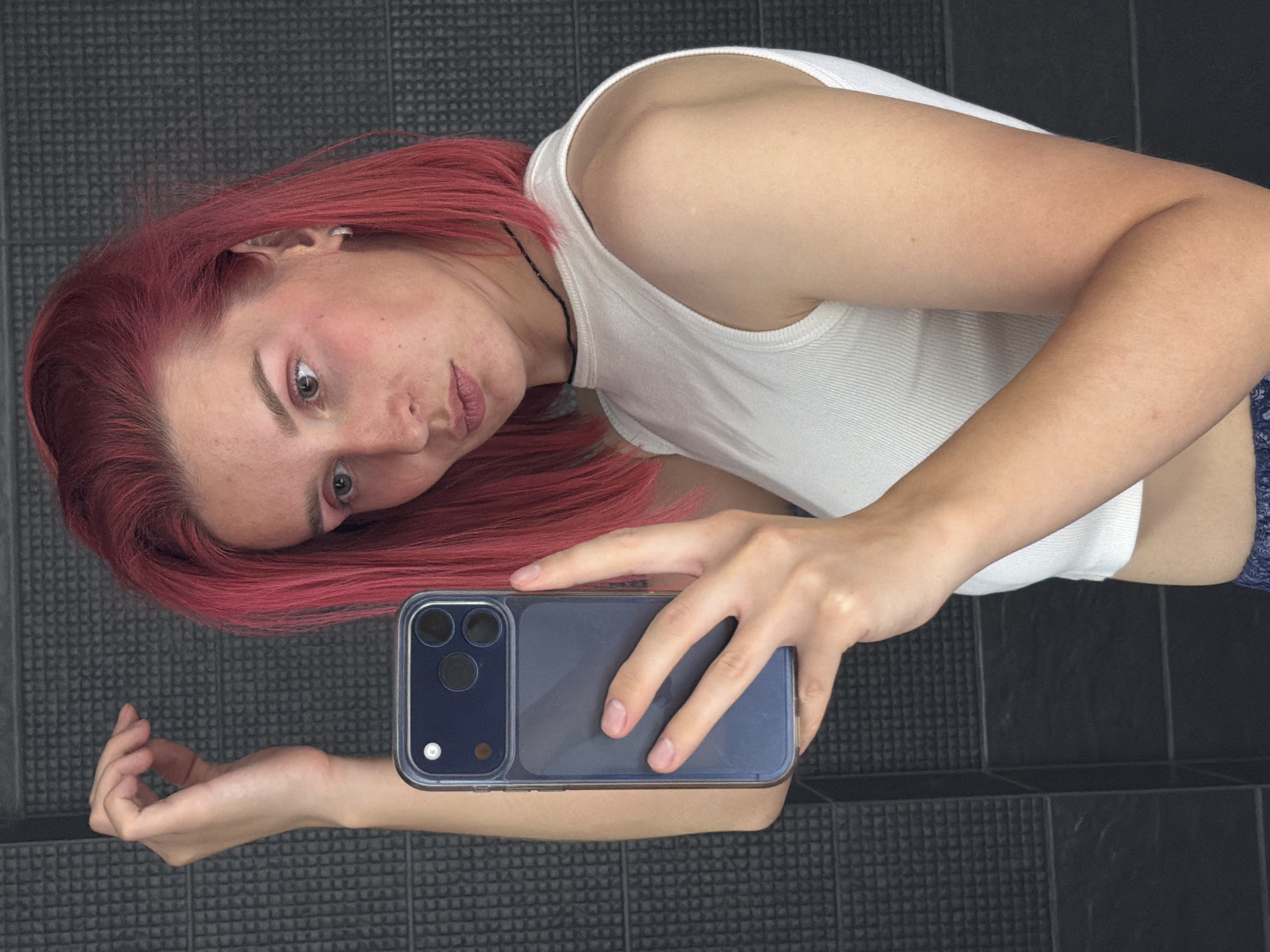 A young person with bright pink hair and makeup is taking a selfie in a bathroom. They are holding a smartphone in their right hand, which is positioned to capture their reflection. The background consists of dark, textured tiles on the wall. The person is wearing a white tank top and blue patterned bottoms. Their left hand is raised above their head. The image appears to be taken in a private residence, likely a bathroom, given the tiled walls and the presence of a mirror for taking a selfie. There is no indication of the specific location within Chaikovsky, Russia, other than the fact that the user has specified the image is from that city.