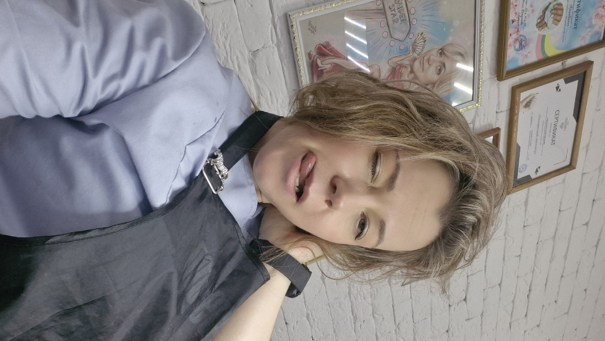 The image features a woman taking a close-up selfie or portrait in what appears to be a professional setting, possibly a beauty salon, against a white brick wall. She has light brown or blonde, medium-length hair and is looking towards the camera with a playful expression, her mouth slightly open and tongue peeking out. Her head is tilted to the right, and her left arm/hand is resting near her head, showing a black smartwatch or wristband.
She is wearing a light blue long-sleeved top or shirt, covered by a black apron. A silver-colored, possibly rhinestone-studded, brooch shaped like a horse or unicorn is pinned to the strap of her apron.
The background consists of a white-painted brick wall, adorned with several framed items. A prominent large frame displays an illustration of a blonde woman with a halo, with the text "SUPER STAR" above her head. Below the illustration, various Cyrillic words are visible, including "Ypocota" (Beauty), "Кристина", "Косунова", "Шихова", and a date "30.12.14".
Next to this, there are other framed certificates. One clearly displays "СЕРТИФИКАТ" (Certificate) and names "Кристина Кайтуристиа" (Kristina Kayturistia) and the skill "Аппаратный маникюр" (Hardware manicure), suggesting the woman might be a certified nail technician. Another smaller framed certificate with the "SEREBRO" logo is also visible. The overall setting implies a professional, likely beauty-related, environment.