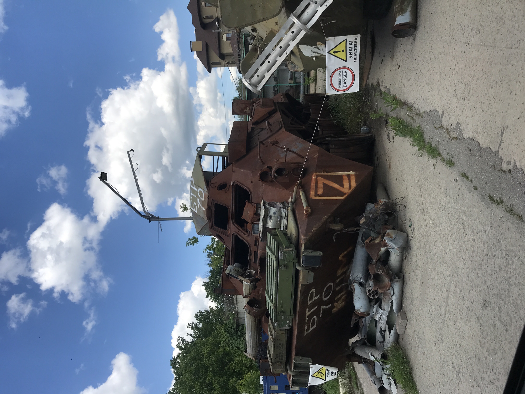 The image displays a heavily damaged and rusted armored personnel carrier (APC), likely a BTR-70, positioned outdoors under a bright blue sky dotted with white clouds. The vehicle shows extensive battle damage, with parts of its structure missing or severely deformed, and its interior exposed. White Cyrillic text "BTP 70" is visible on its side, along with a prominent orange "Z" symbol, often associated with Russian military forces.
Beside the APC, a pile of metallic debris, appearing to be fragments of shells or other ordnance, rests on a concrete surface. In the background, other damaged military equipment, possibly a tank or another armored vehicle, can be partially seen, along with a building featuring a reddish-brown roof and green trees. The ground is a mix of concrete and grassy patches.
Several warning signs are present: a yellow triangular sign with an exclamation mark reads "УВАГА! НЕБЕЗПЕКА!" (Attention! Danger!), and a red circular sign beneath it reiterates "НЕБЕЗПЕКА DANGER" in both Cyrillic and English. Other faint Cyrillic text, such as "KUIM," and numbers like "4345" and "350" are visible on the vehicle and in the background.
The scene depicts a static display of war-damaged equipment, likely an exhibit or memorial showcasing the aftermath of conflict, possibly in Ukraine given the visible Cyrillic text and symbols. The time is daytime, characterized by bright sunlight.