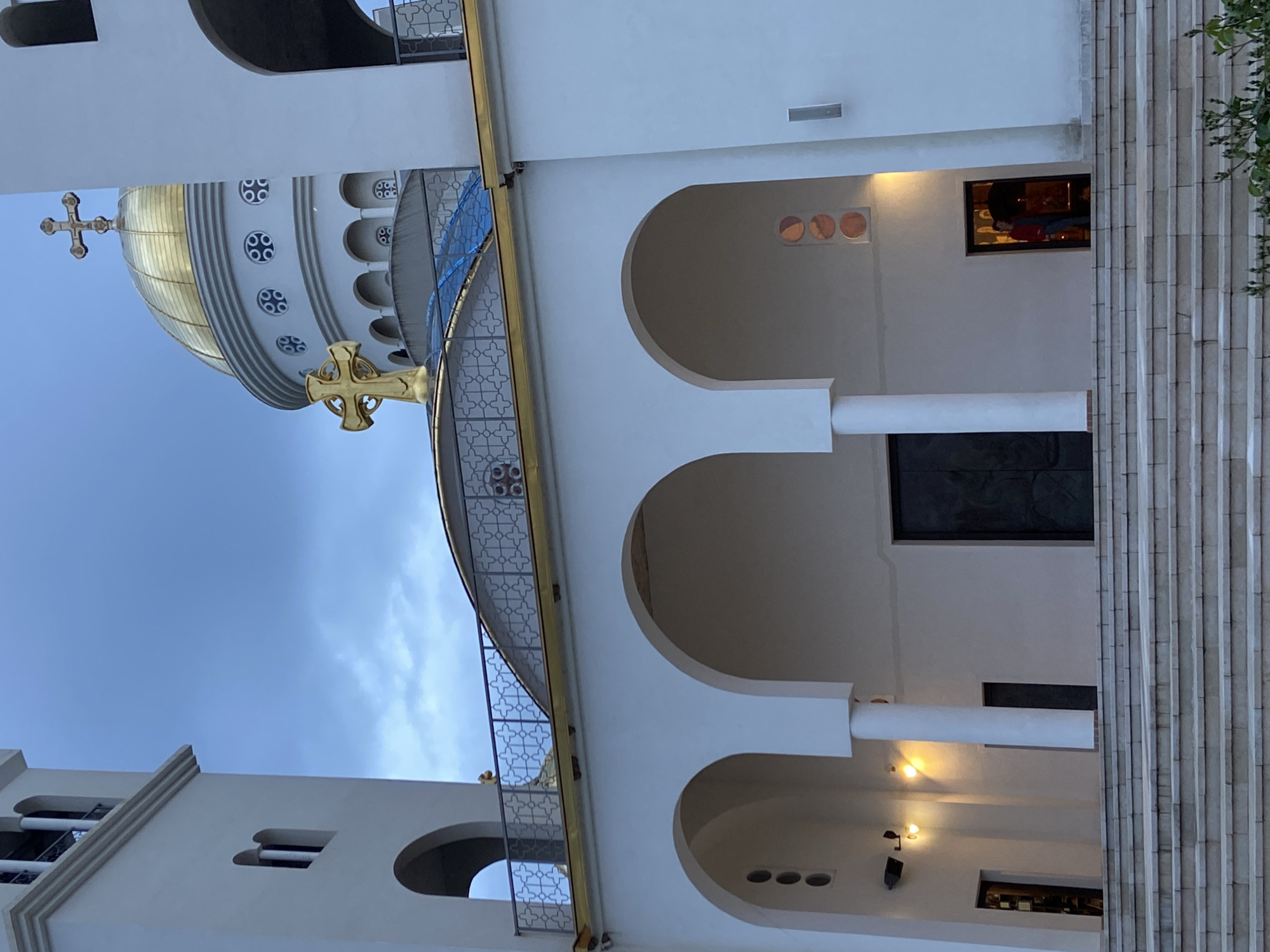 The image captures a grand, white architectural structure, identified as an Eastern Orthodox cathedral, likely the Cathedral of Saint Jovan Vladimir in Bar, Montenegro. The scene is set during daylight hours, characterized by a light blue sky with wispy clouds.
The most striking features are two prominent golden domes, each crowned with a distinctive golden Orthodox cross. The larger dome in the upper left section of the image showcases intricate detailing with circular windows arranged around its base. The building itself is predominantly white, featuring a series of gracefully curved arches and sturdy pillars that create a sense of depth and grandeur. Decorative metal railings with repeating patterns adorn balconies and walkways.
On the right side of the composition, a wide, multi-tiered staircase ascends, flanked by textured vertical columns. Several interior light fixtures are visible through arched openings and windows, emitting a warm, subtle glow that contrasts with the bright exterior. A rectangular window on an upper level offers a partial glimpse into an interior space, where a dark, indistinct form or object can be discerned. The overall impression is one of architectural majesty and spiritual significance.
No people are visible, and there is no discernible text within the image. The building appears well-maintained, suggesting a place of ongoing worship or cultural importance.