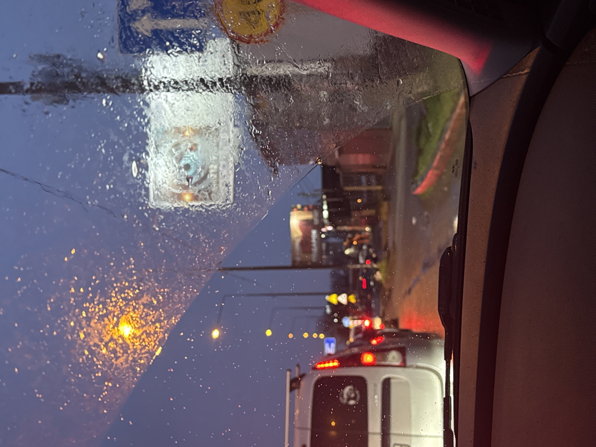 The image depicts an urban street scene at dusk or night, viewed from the interior of a vehicle through a rain-splattered windshield. The foreground shows the vehicle's dark dashboard and a red-tinged right A-pillar. A windshield wiper is visible at the base of the windshield.
Outside, a multi-lane road is wet, reflecting ambient lights. Traffic is present and appears to be stationary or moving slowly. Immediately ahead of the viewer's vehicle is the rear of a white utility van, possibly a Ford Transit Custom, with its red taillights illuminated. The van features a roof rack and a partially visible license plate ending in "B". Behind the van, other vehicles with illuminated red taillights form a line of traffic.
Streetlights emit orange-yellow light, visible as blurred orbs through the rain droplets. Several road signs are discernible, including a blue rectangular sign with a white upward-pointing arrow, a yellow triangular warning sign with two curving black arrows, and a circular red-bordered sign. On the right, a large, illuminated billboard displays an image resembling an eye. Further down the street, blurred commercial buildings and additional illuminated signs are visible. No individuals are clearly identifiable. The location is an unnamed city, province of Unknown, Unknown.