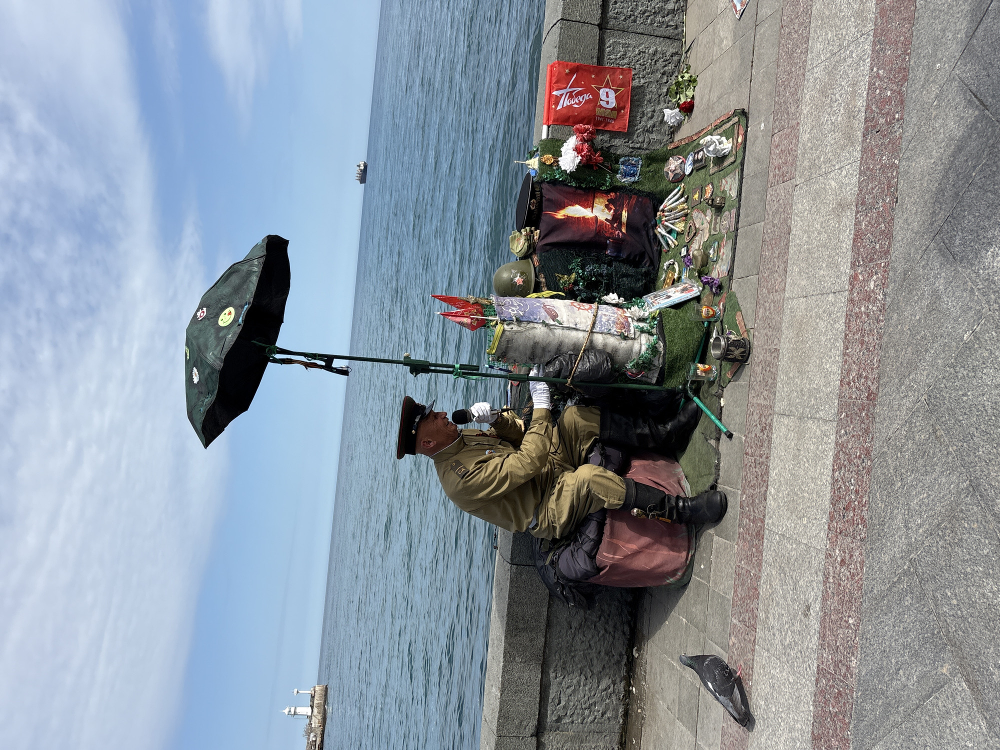 On a sunny day in город Ялта, Ukraine, a man in a military uniform is sitting on a suitcase by the sea. He is holding a microphone and appears to be singing. An umbrella is propped up to shield him from the sun. Behind him is a display of flags, flowers, and other memorabilia, including a flag with the number 9 and the words "Победа" (Victory) and "Май 1941-1945" (May 1941-1945). A pigeon is on the ground nearby. In the background, the sea stretches to the horizon, with a ship visible in the distance. A lighthouse can be seen on the left. The man is sitting on a paved walkway next to a stone wall.