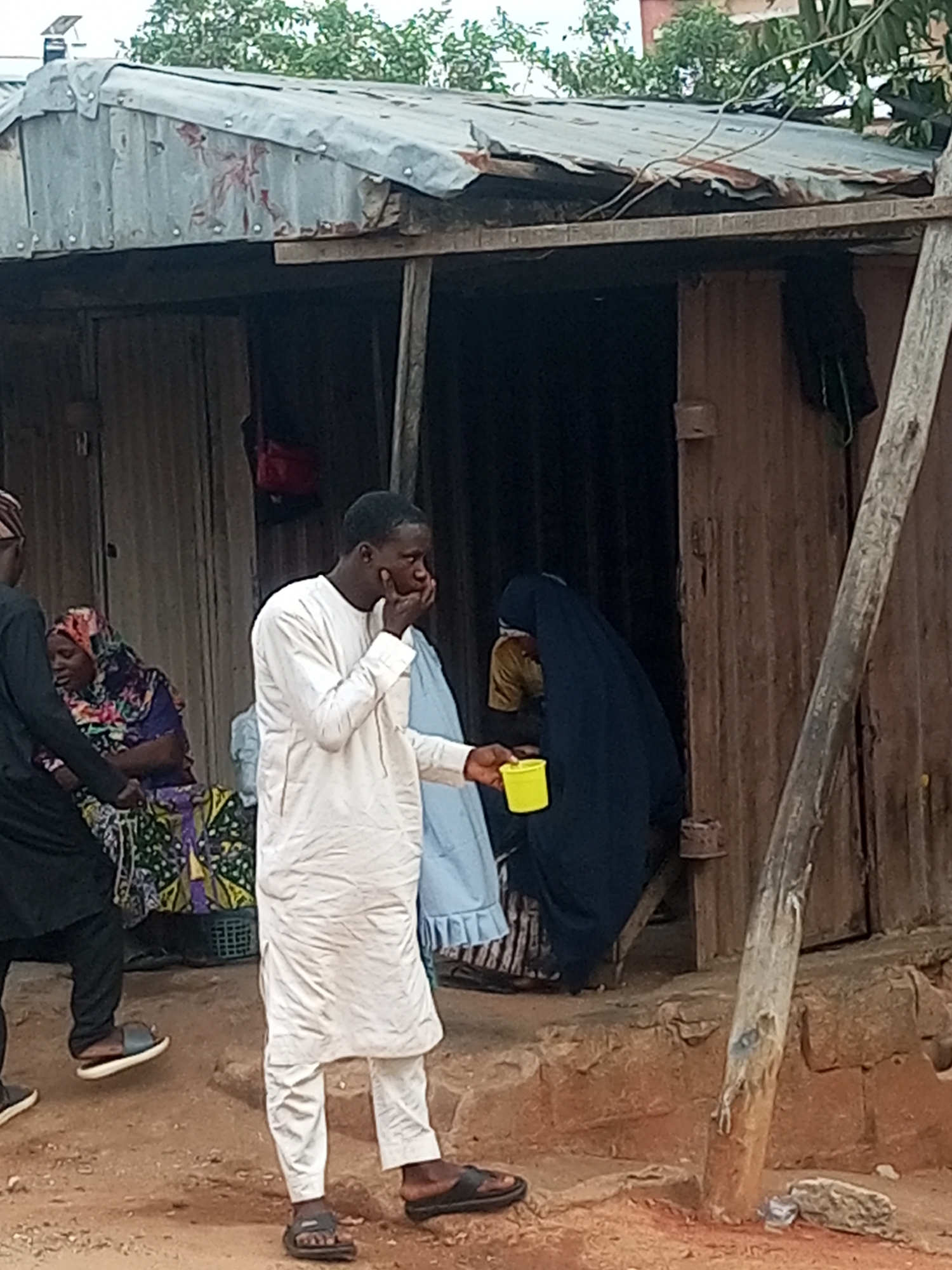 In Garko, Nigeria, a man in a white traditional outfit stands in front of a dilapidated structure with a corrugated metal roof. He is holding a yellow cup in his right hand and appears to be speaking. Behind him, a woman in a dark hijab and a yellow garment is partially visible. To the left, another woman wearing a colorful headscarf and patterned fabric sits near a person dressed in black. The ground is uneven and appears to be made of dirt and rocks, with some vegetation visible in the background. The structure itself is rustic, with wooden beams and corrugated metal walls.