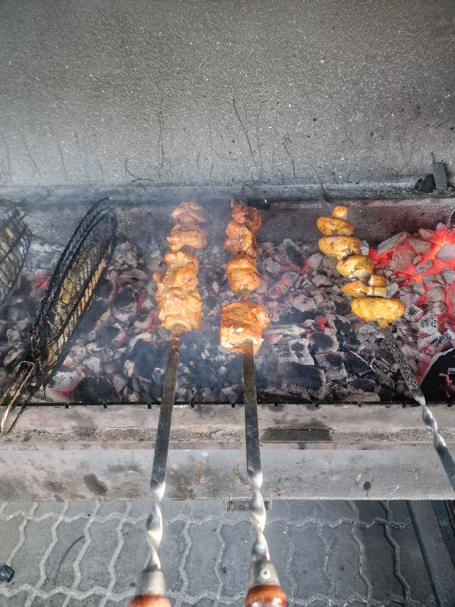A close-up, overhead shot shows food being grilled on a charcoal barbecue in Zelenogradsk, Russia. The barbecue contains glowing red coals and ash. On the left, a fish is grilled inside a black metal cage. To the right of the fish, there are two skewers of chicken pieces, and further to the right, a skewer of mushrooms. Smoke is rising from the barbecue. The background consists of a grey, textured wall, and the foreground shows a patterned grey pavement.
