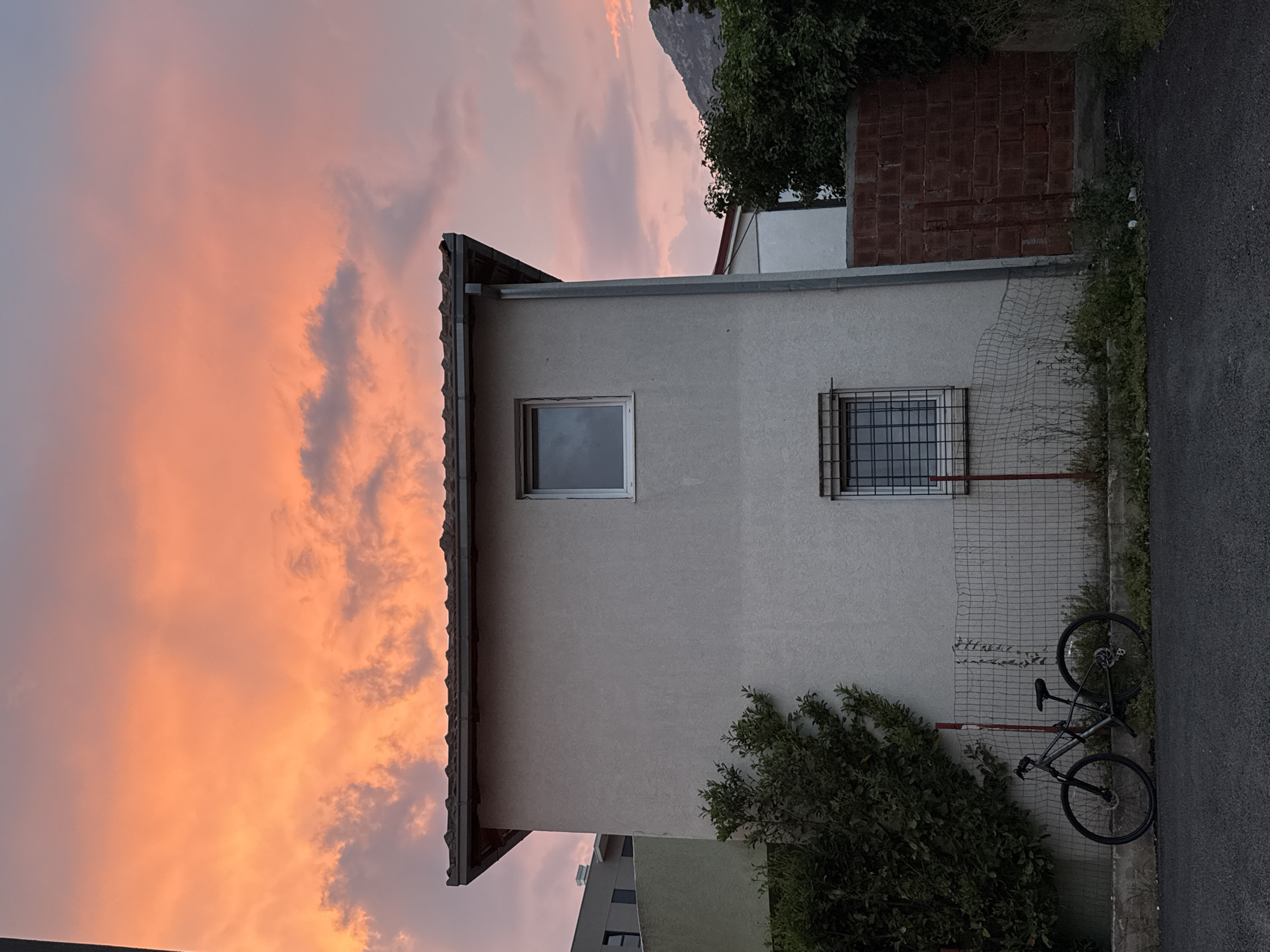 A two-story building with a stucco facade and a tiled roof stands under a dramatic sky filled with vibrant orange and pink clouds during sunset. The building has a simple design with two visible windows, one on the upper floor and another on the lower floor that is protected by a metal grate. To the left of the building, a dark green tree with dense foliage grows. In front of the building, a bicycle with a dark frame is parked near a wire fence. The ground in front of the fence is covered with dry grass and weeds, and a paved surface is visible in the foreground. To the right of the building, a section of red brick wall is partially visible, and more trees with dark green leaves complete the scene. In the distance, a dimly lit mountain range can be seen. The overall impression is of a residential area in the city of Bar, Montenegro, captured at dusk with an impressive celestial display.