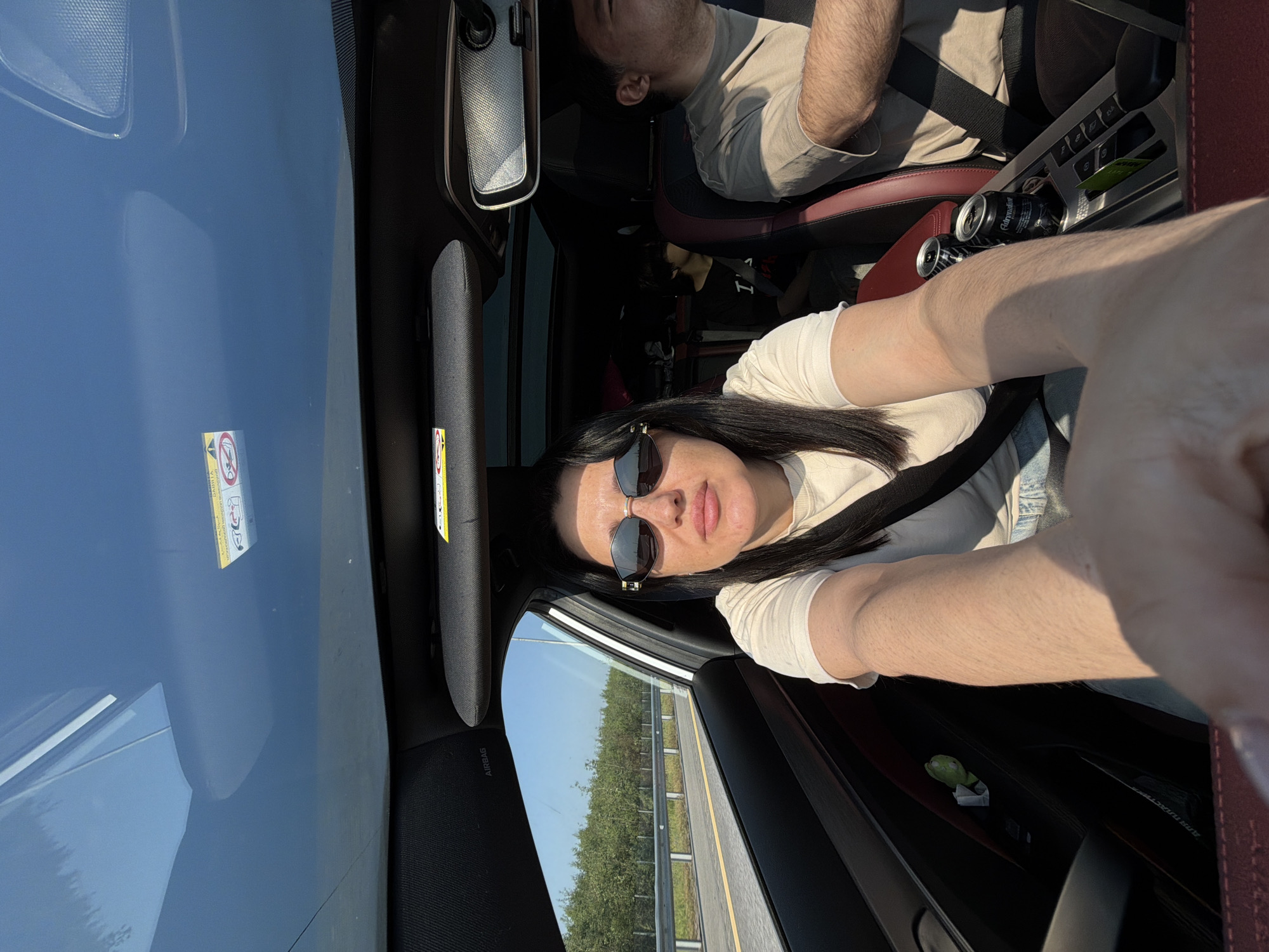 A woman is driving a car on a sunny day. She is wearing sunglasses and a light-colored t-shirt. The interior of the car is visible, including the dashboard, seats, and a panoramic sunroof. Outside the car, trees and a highway are visible. There are two cans of soda in the center console of the car, and a man is in the passenger seat. A child is visible in the back seat. The image appears to have been taken in the city of Fyodorovo, Russia.