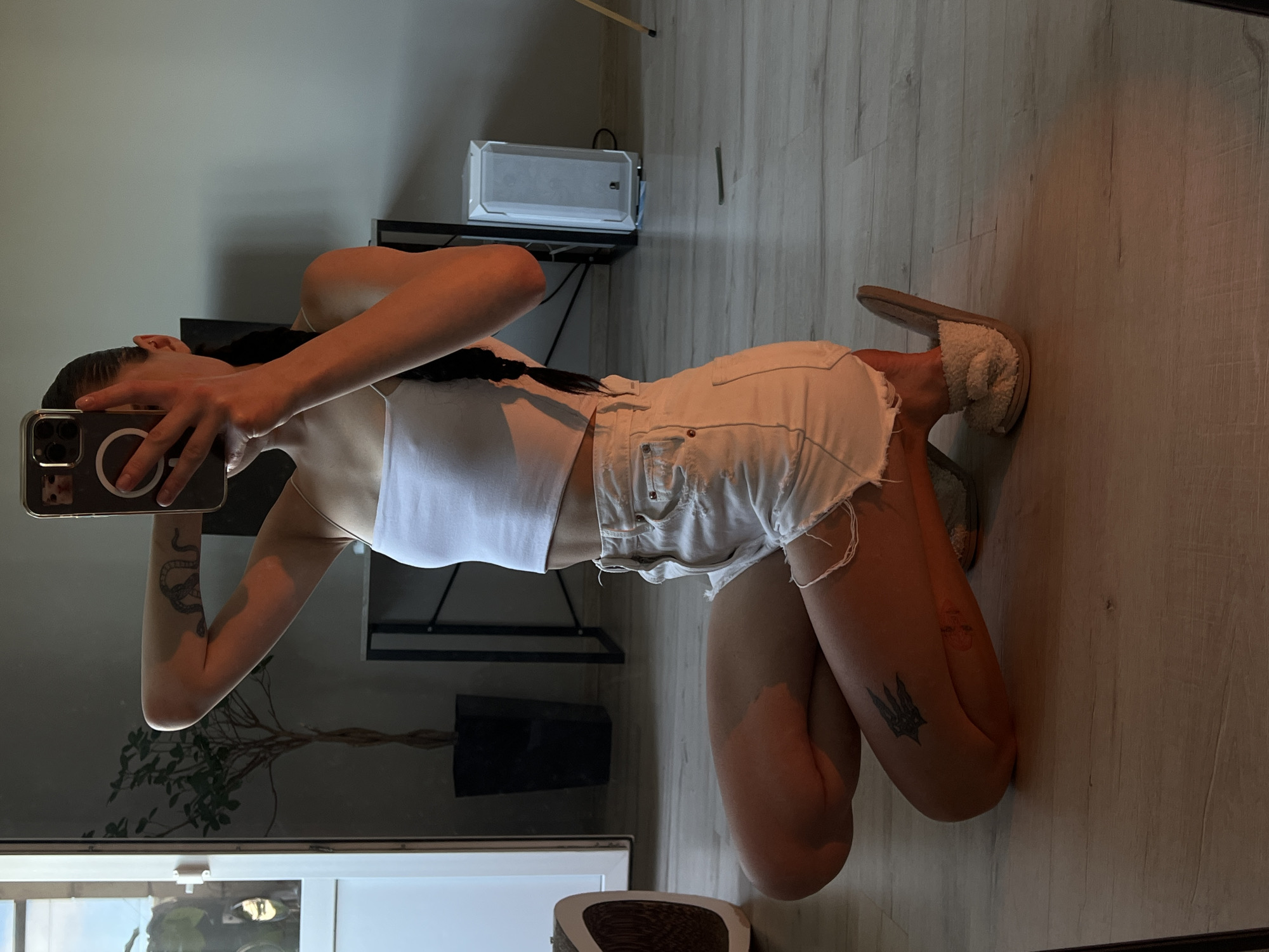 A woman is kneeling on a wooden floor, taking a selfie in a mirror. She is wearing a white tank top and white denim shorts. She has a tattoo on her right arm and another on her left thigh. The background shows a modern apartment with a plant, a desk, and a computer tower. The image was taken in Ternopil, Ukraine.