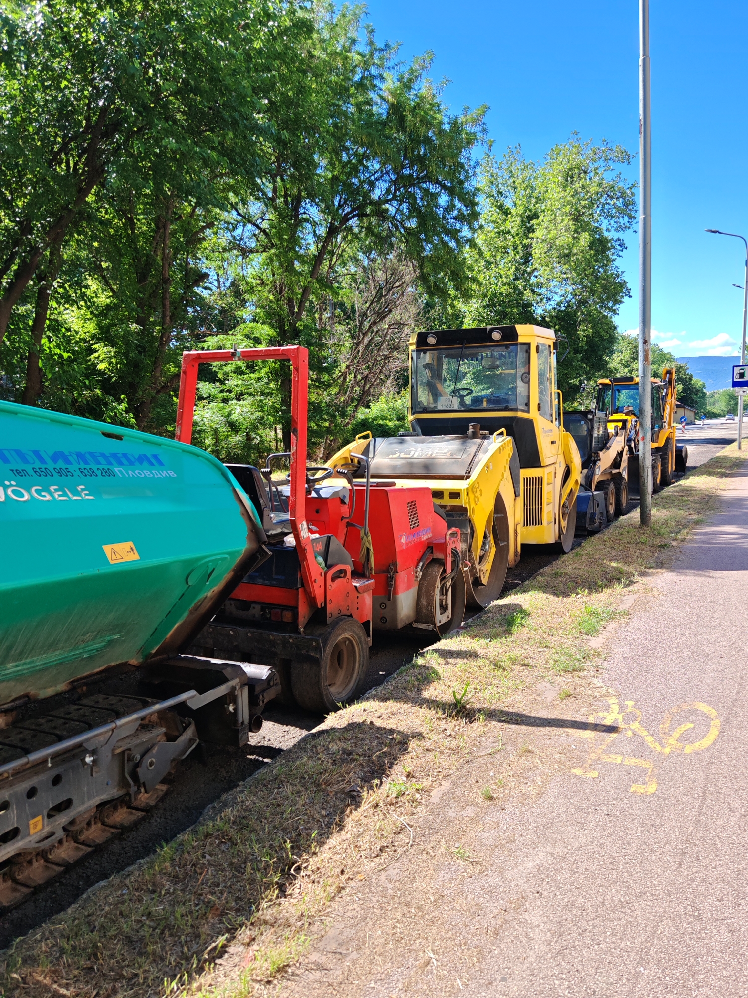 This image depicts a street paving scene in Plovdiv, Bulgaria. In the foreground, a green asphalt paver, identified with Bulgarian text including "тел. 650 905", "638 280", and "ПЛОВДИВ", and "VÖGELE", is parked on the side of a road. Behind it, a red and black paving machine with rollers is attached. Further down the road, a line of yellow construction vehicles, including a road roller and a backhoe loader, are parked. The background features a line of green trees and a bright blue sky, suggesting it is a sunny day. The road itself has yellow markings, including a bicycle symbol on the asphalt. The scene implies ongoing road construction or maintenance.