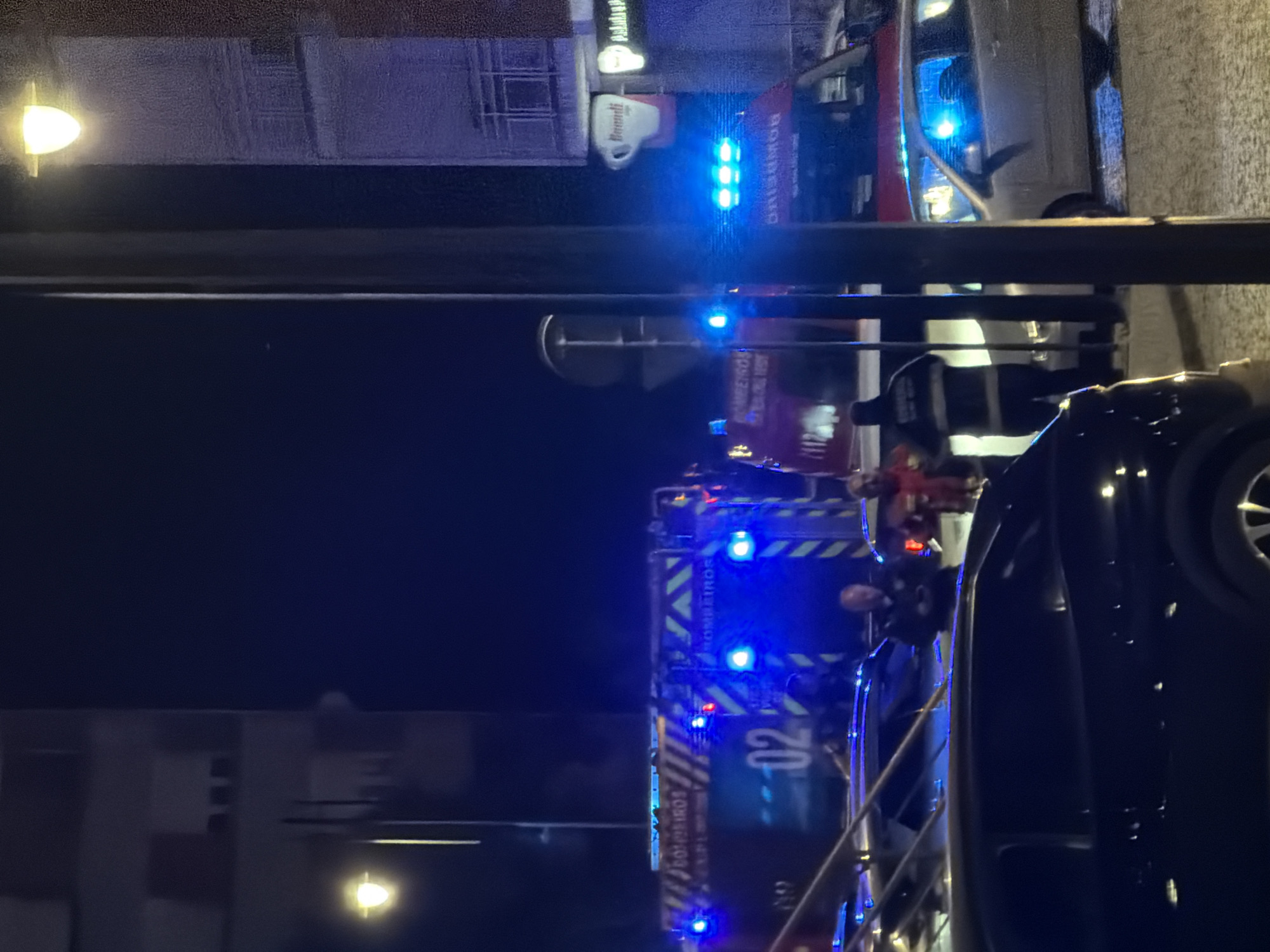 At night in Vila Real, Portugal, a fire truck labeled "BOMBEIROS" with the number "02" is parked on the street. Emergency personnel are gathered around the vehicle. A dark-colored car is parked on the right side of the frame. Buildings are visible in the background. The scene is illuminated by streetlights and the flashing blue lights of the emergency vehicles. A store sign that reads "Danutti" is visible in the background.