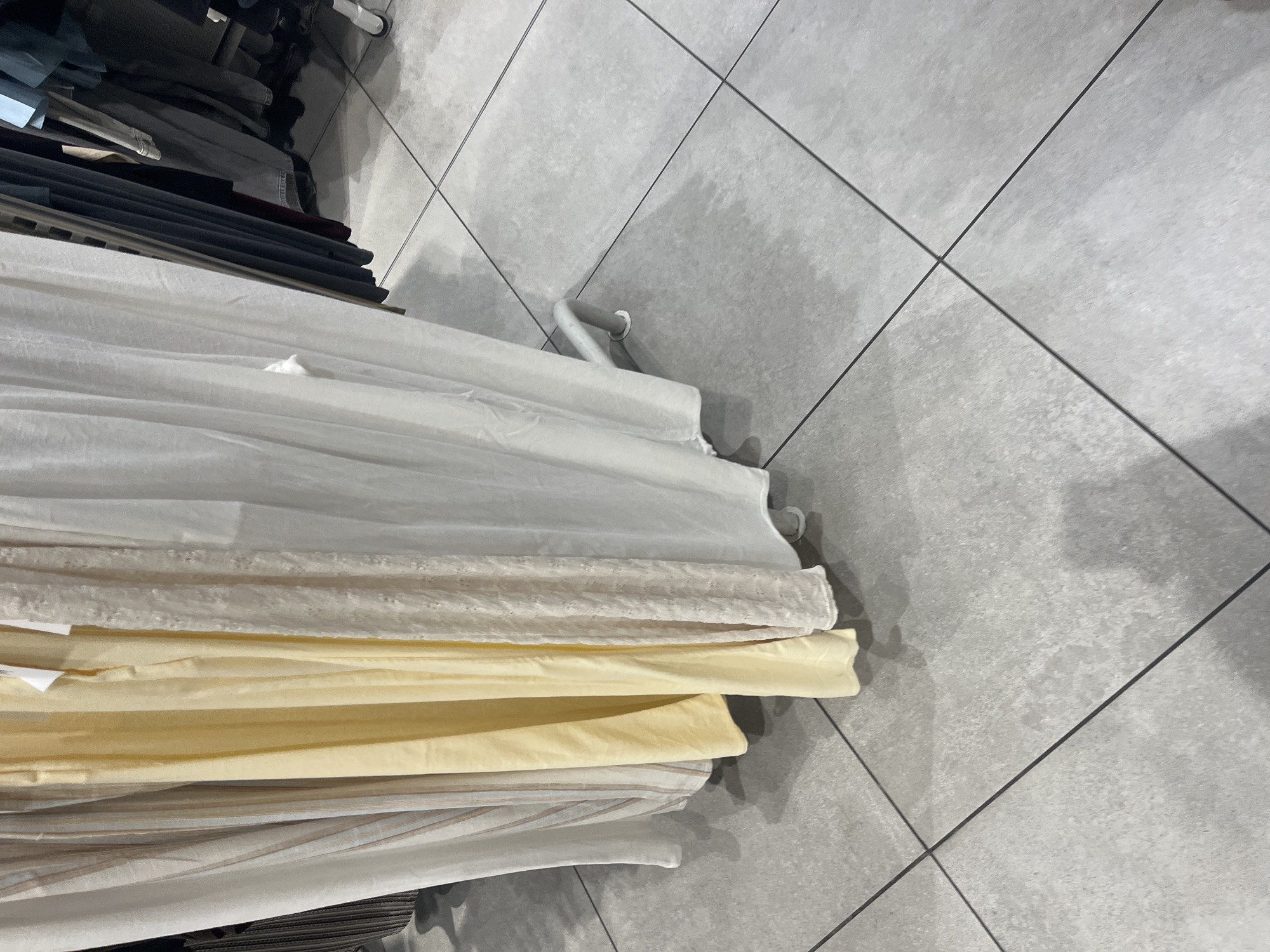 A rack of folded fabrics, predominantly white and pale yellow, sits on a gray tiled floor. The rack is partially visible, with some clothes hanging above the fabrics. The location is not identifiable as Blagoevgrad, Bulgaria, based on the visual information provided.