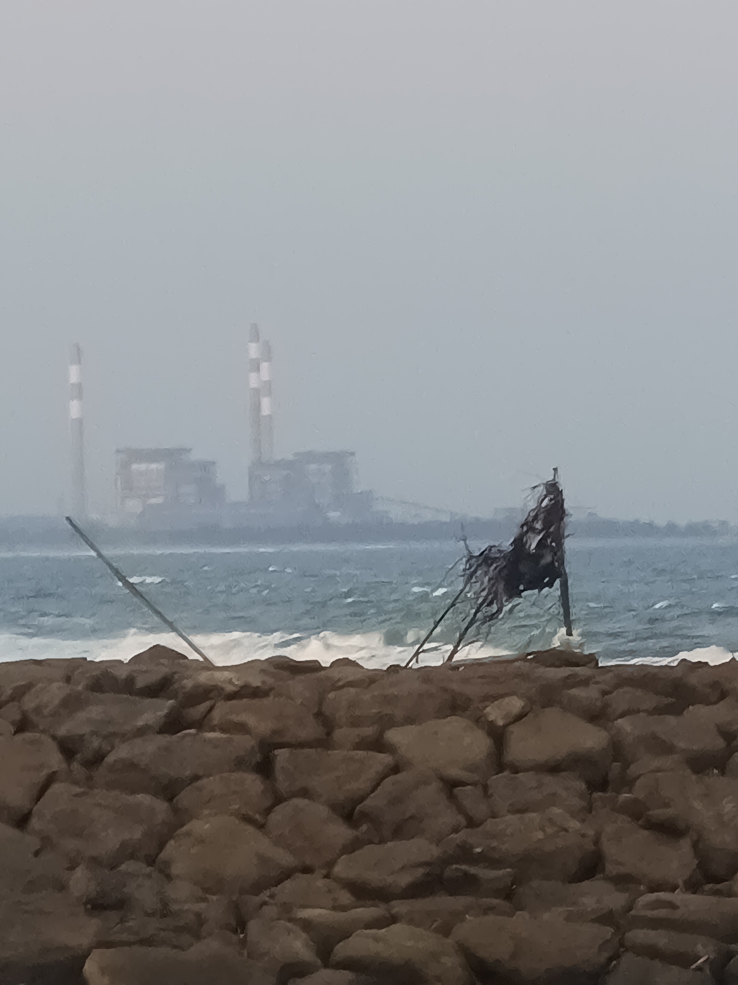 The image captures a somewhat hazy coastal scene in the city of Kelurahan Tegalkamulyan, Cilacap, Indonesia. In the background, a large industrial complex with tall, striped smokestacks is visible through the mist. In the middle ground, the ocean churns with white-capped waves. In the foreground, a breakwater constructed of rough, dark brown rocks is prominent. To the right of the frame, a tattered black flag or piece of debris is attached to a makeshift pole, appearing to be caught by the wind and waves. Two slender poles, possibly part of a pier or temporary structure, extend diagonally into the water from the left side of the frame. The overall atmosphere is muted and overcast.