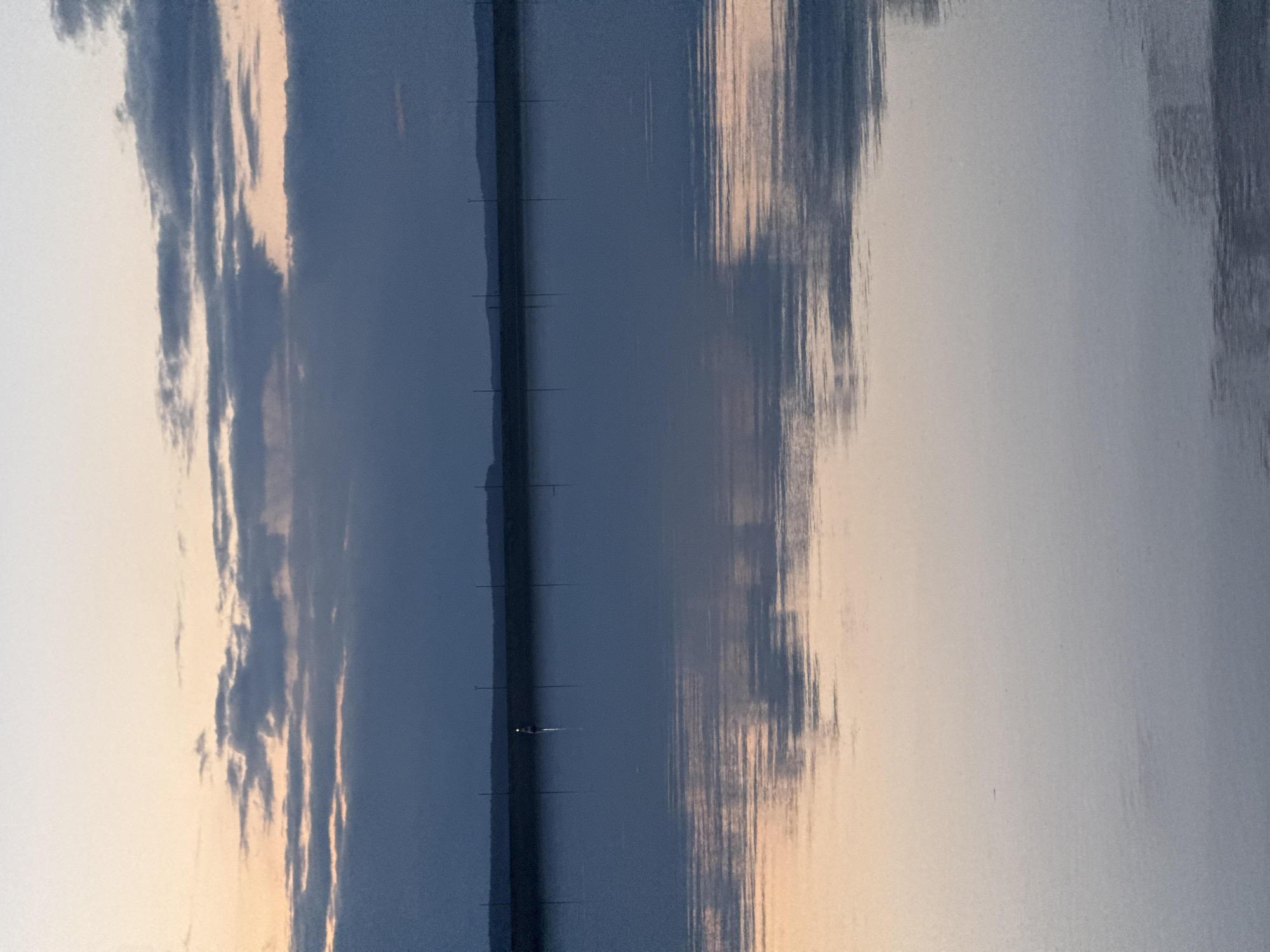 A serene landscape, likely captured at dawn or dusk, depicts a wide body of water beneath a partially cloudy sky. The image, although oriented vertically, captures a horizontal scene. The upper half features a cloudy sky, with light gray-blue clouds contrasting with softer, light orange and pink hues, indicating indirect sunlight.
A narrow, dark horizontal strip stretches across the middle of the image, separating the sky from its reflection. This strip appears to be a distant landmass or a low-lying structure, upon which several faint, short vertical lines, possibly utility poles or navigation markers, are visible. Near the center of this strip, a small, bright white speck, likely a distant boat with a light or a buoy, stands out.
The lower half of the image is dominated by the calm water, which beautifully reflects the sky and clouds above. These reflections are slightly elongated and appear as soft, vertical streaks of color, mirroring the blues, grays, and warm orange-pink tones of the sky.
The overall color palette is muted, with cool blues and grays predominating, complemented by gentle warm tones from the diffused light. The scene evokes a tranquil and contemplative mood. The image displays some visual noise or grain, suggesting it was captured in low light conditions. There are no identifiable people, explicit activities, or visible text.