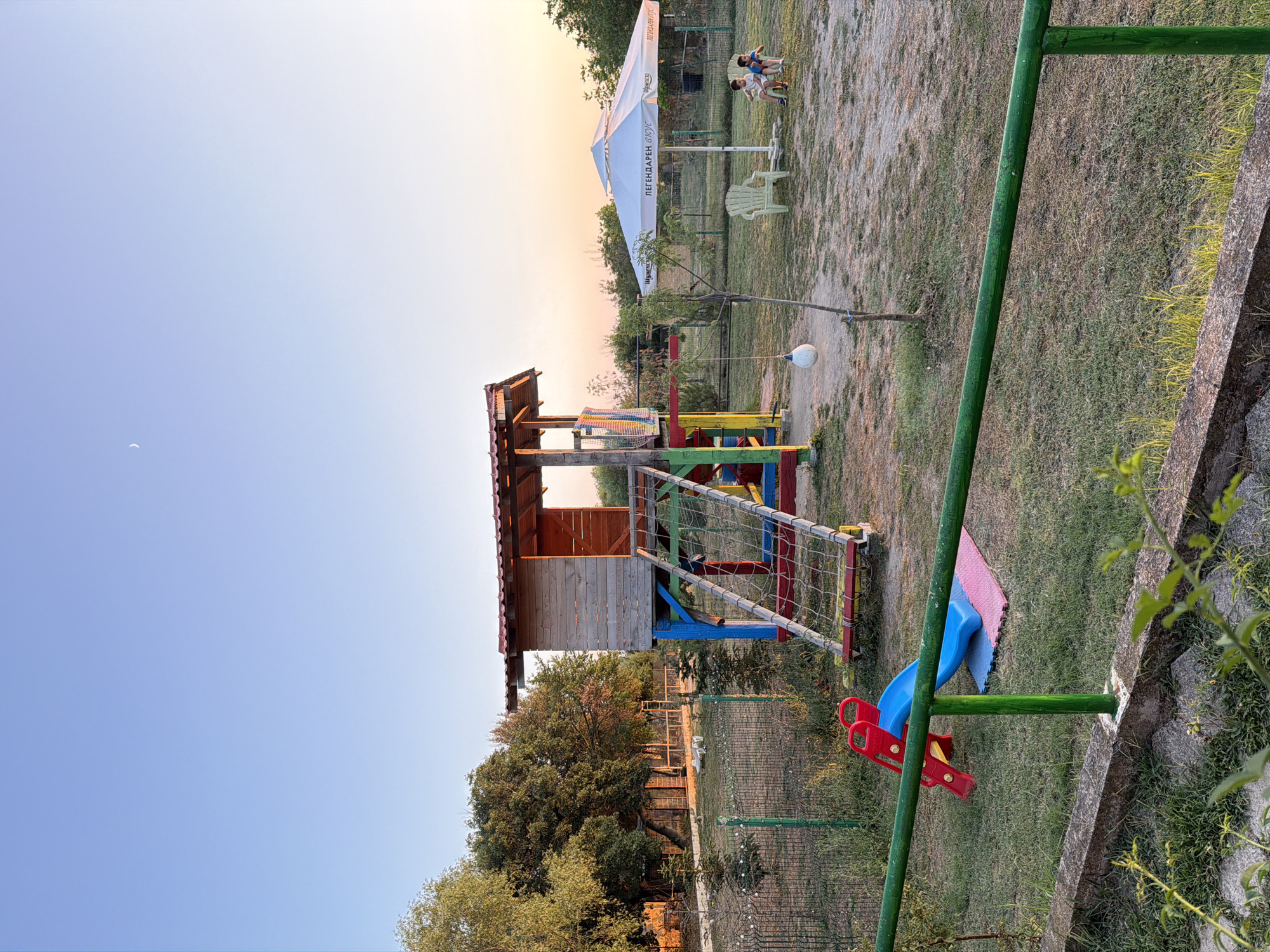 The image depicts an outdoor children's playground at dusk, possibly located in Tvarditsa, Bulgaria, given the context of the prompt. The playground features a wooden playhouse elevated on colorful stilts, with a rope ladder providing access. A red and blue slide is situated in the foreground, resting on a pink and blue mat. A green metal railing runs horizontally across the lower part of the image, separating the playground from a grassy area. In the background, two young boys are seated on chairs under a large white umbrella. The sky is a clear, pale blue with a faint crescent moon visible. The overall environment appears to be a park or garden, with trees and vegetation surrounding the play area.