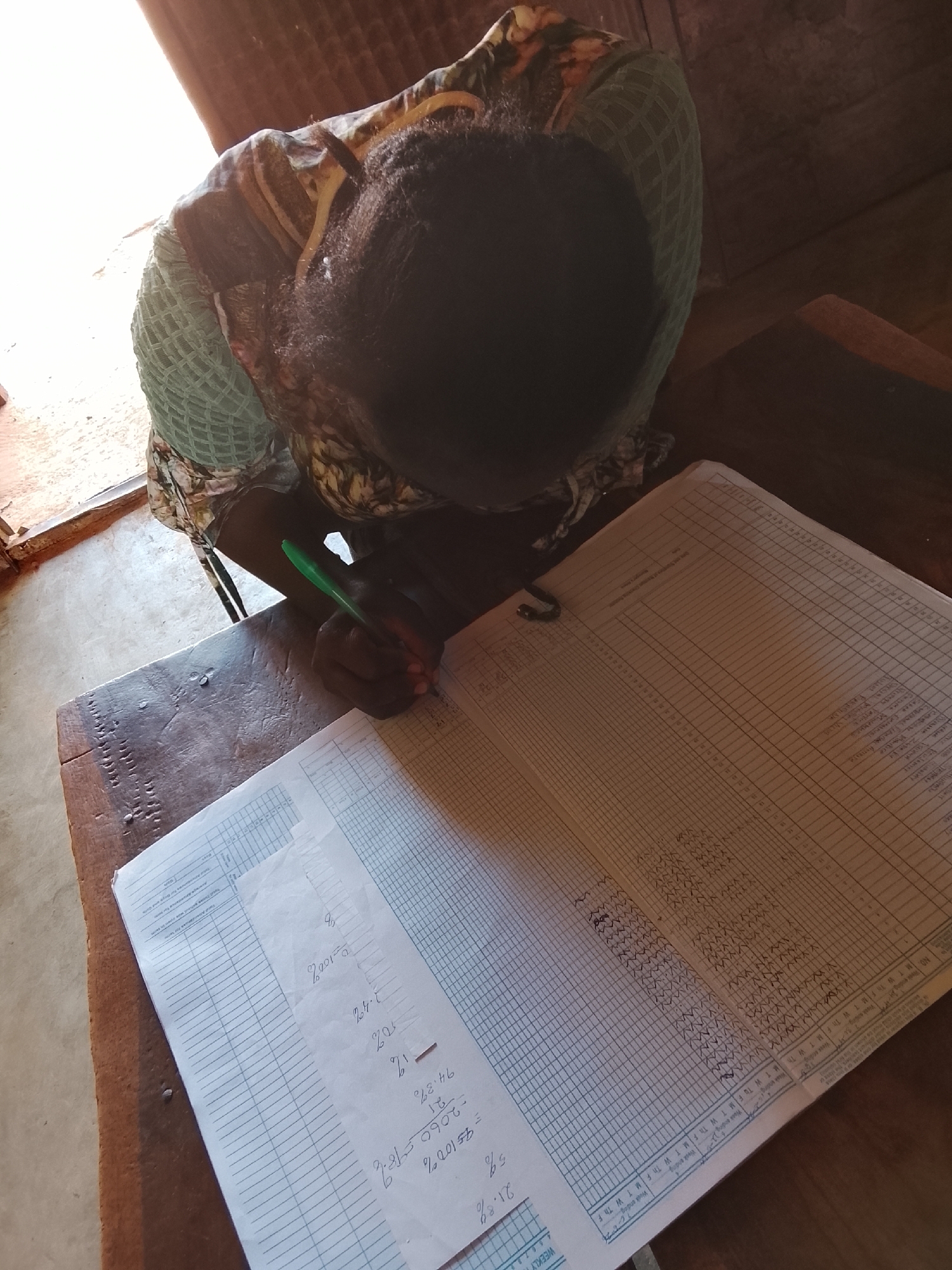 A woman with dark hair is seated at a worn wooden table, intently writing in a large, lined ledger with a green pen. The ledger, filled with handwritten entries and a grid-like chart, lies open before her. A separate sheet of paper, displaying handwritten calculations involving percentages, rests on top of the ledger. The scene is bathed in natural light, likely from a nearby window, suggesting an indoor environment. This appears to be a basic office space; it cannot be confirmed that this photo was taken in Akwanga, Nigeria.