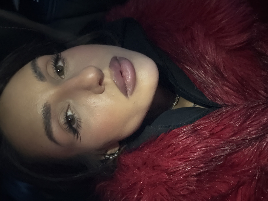 An adult female is captured in a close-up, chest-up portrait, looking directly at the viewer. She has dark hair, styled away from her face, and brown eyes accentuated by prominent winged eyeliner and long, dark eyelashes. Her eyebrows are dark and well-defined, and her lips are full, glossy, and light pink. She wears a voluminous, deep red textured faux fur jacket over a dark garment, possibly a hooded top. A thin, gold-colored chain necklace is visible around her neck, and a silver-colored, possibly hoop, earring adorns her left ear. The background is uniformly dark and featureless. The scene is situated in Moscow, Russia.