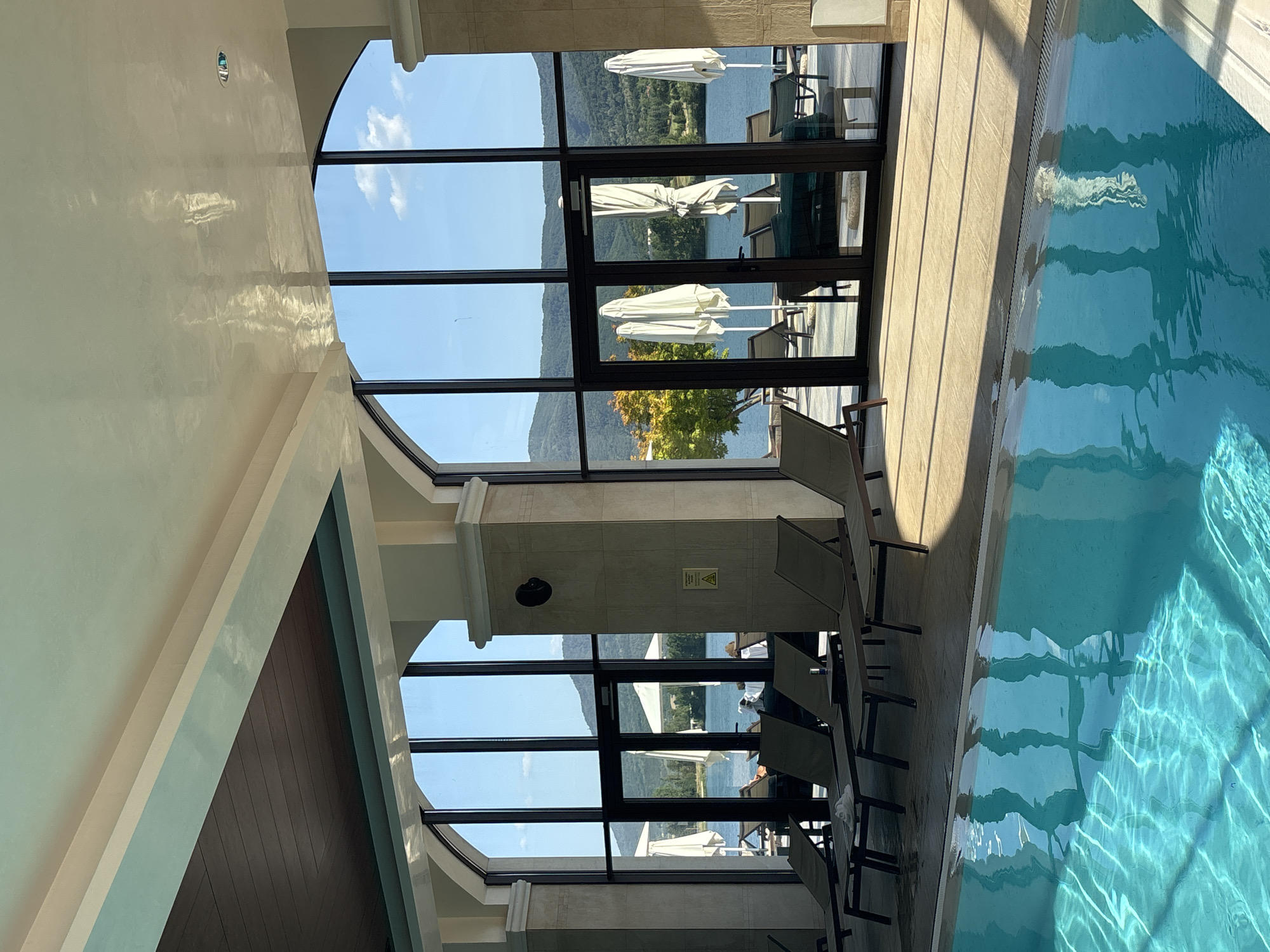 This image depicts an indoor swimming pool area with a view of a lake and surrounding hills. The pool's turquoise water occupies the foreground, with sunlight reflecting on its surface. Beyond the pool, several lounge chairs are arranged on a tiled patio. Large arched windows and glass doors offer a panoramic view of the outdoor scenery, which includes the lake, trees with autumn foliage, and distant mountains under a clear blue sky with scattered clouds. Several white umbrellas are visible outside. The architecture of the indoor space features high ceilings with a decorative finish and wooden accents. The setting is identified as being in the city of Razliv, Bulgaria, likely a resort or hotel due to the amenities visible. There are no people clearly engaged in any activity within the frame, though some figures are faintly visible through the glass doors outdoors.
