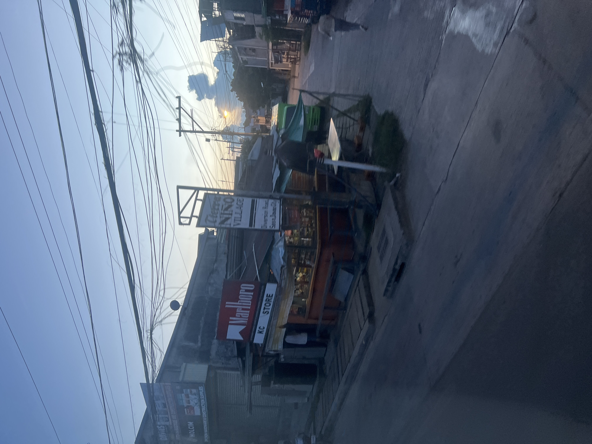 The image captures a street scene at dusk in what appears to be a commercial area of Davao City, Philippines. A prominent Marlboro sign is visible above a store labeled "KC STORE," suggesting it's a small convenience store or sari-sari store. Next to it, a sign reads "Tierra NINO VILLAGE" with an address indicating "Diversion Road Sasa, Davao City." This provides a specific geographical context.
The street is paved, with some areas showing reflections, possibly from recent rain. Utility wires crisscross the sky, a common sight in Philippine urban landscapes. In the background, there are buildings of varying sizes and styles, some appearing residential and others commercial. A green truck is parked on the side of the road, and a person is walking away from the camera on the right side of the street. The sky is a blend of dusky blues and oranges, indicating the transition from day to night. The overall impression is of a typical neighborhood setting in Davao City during the early evening hours.