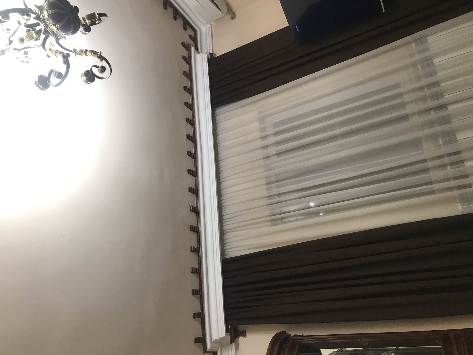 The image shows an interior space, likely a room in a building. A chandelier is partially visible in the upper left corner. The wall is painted a light color. A window covered with blinds and dark brown fabric is present on the right side of the frame. Part of a dark wood structure is visible at the bottom of the image.
There is no information available to determine the location in the city of Yerevan, Armenia.