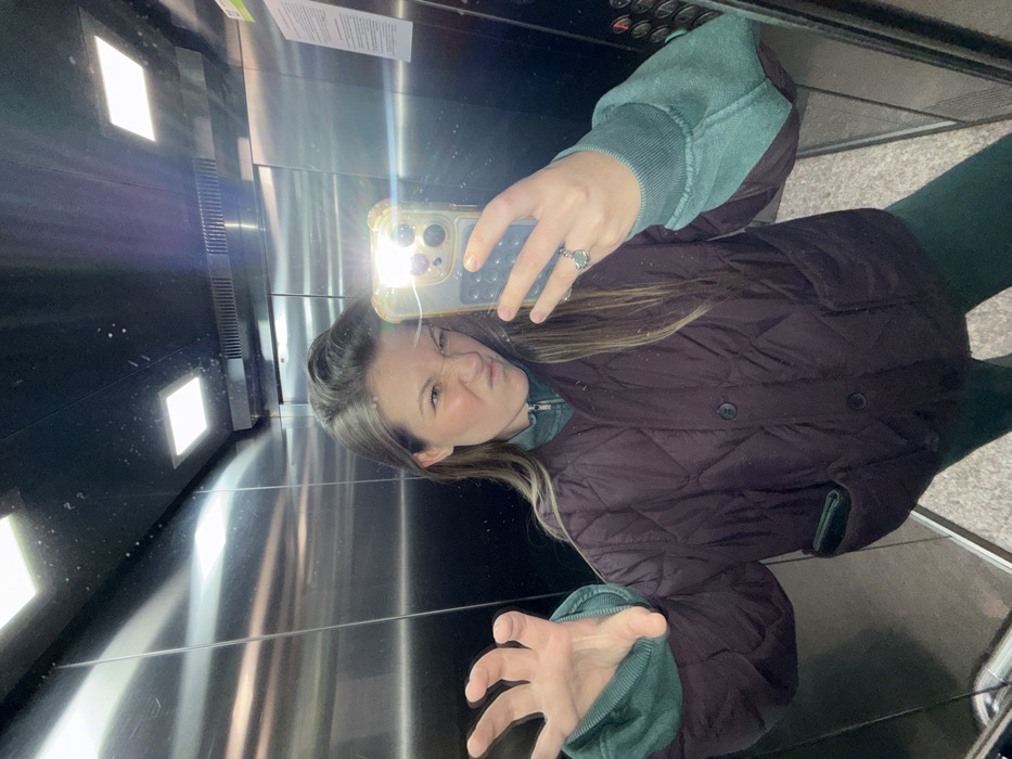The image displays a mirror selfie of a female individual inside an elevator located in Budva, Montenegro. The individual, with long brown hair, is centrally framed, wearing a dark, possibly maroon or deep brown, quilted jacket over a teal or dark green zippered top and matching leggings. Her right hand holds a smartphone, its flash creating a bright glare on the reflective surface; a ring with a light-colored stone is visible on her right ring finger. Her left hand is raised in a claw-like pose, fingers spread. Her facial expression is a playful grimace, with narrowed eyes and a puckered mouth. The elevator interior features polished metallic walls reflecting square ceiling lights, and a control panel with buttons is visible in the reflection on the right side. The floor is a light-colored, hard surface.
