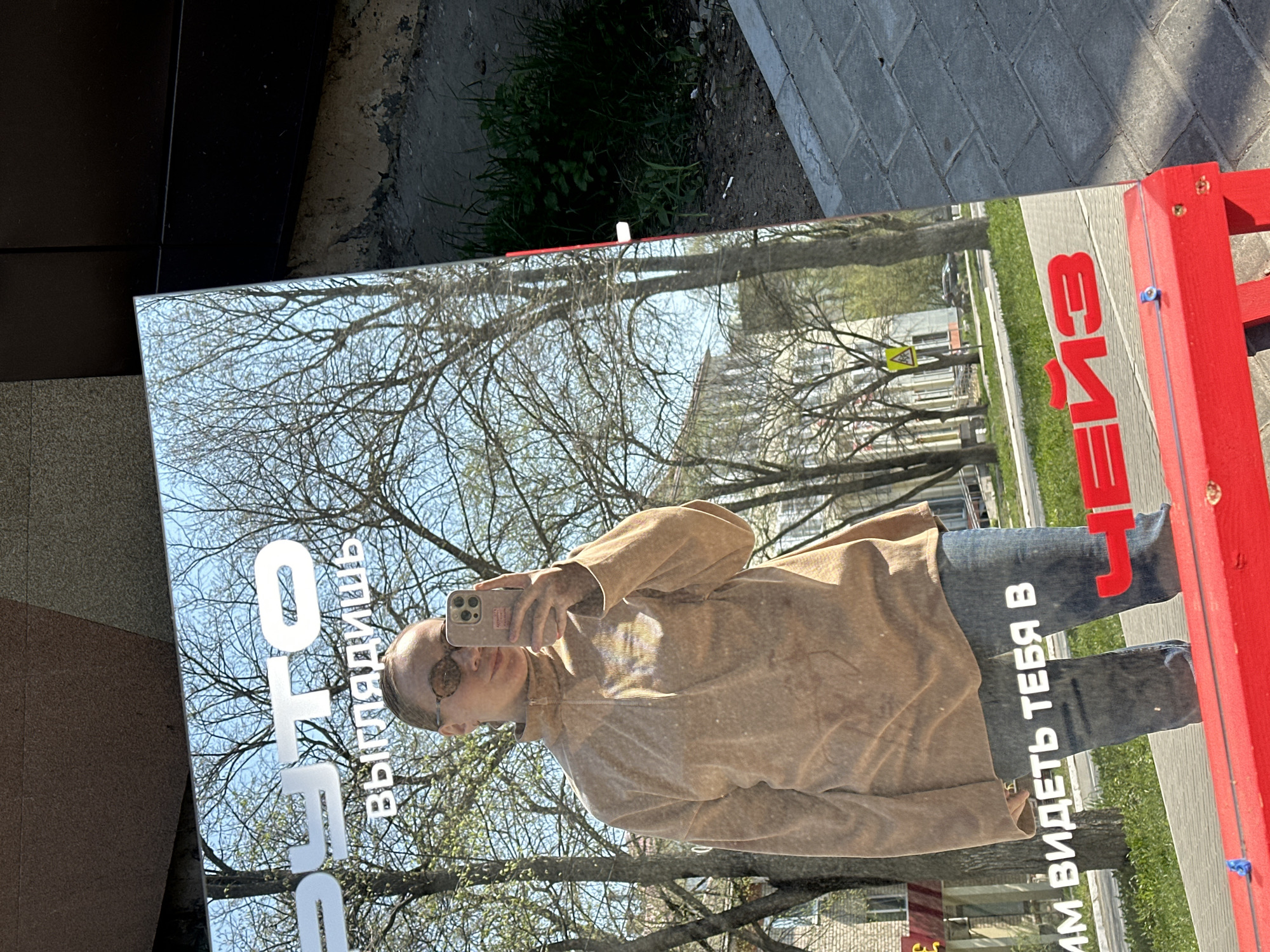 The image shows a mirror positioned on a sidewalk in what appears to be a Russian city, based on the Cyrillic text present. The reflection in the mirror captures a woman taking a selfie with her smartphone. The environment reflected in the mirror includes bare trees, buildings, and what seems to be a pedestrian area. The mirror is mounted on a red frame, which also has Cyrillic writing on it. The setting suggests an urban environment, possibly in the city of Chaikovsky, Russia, considering the language.