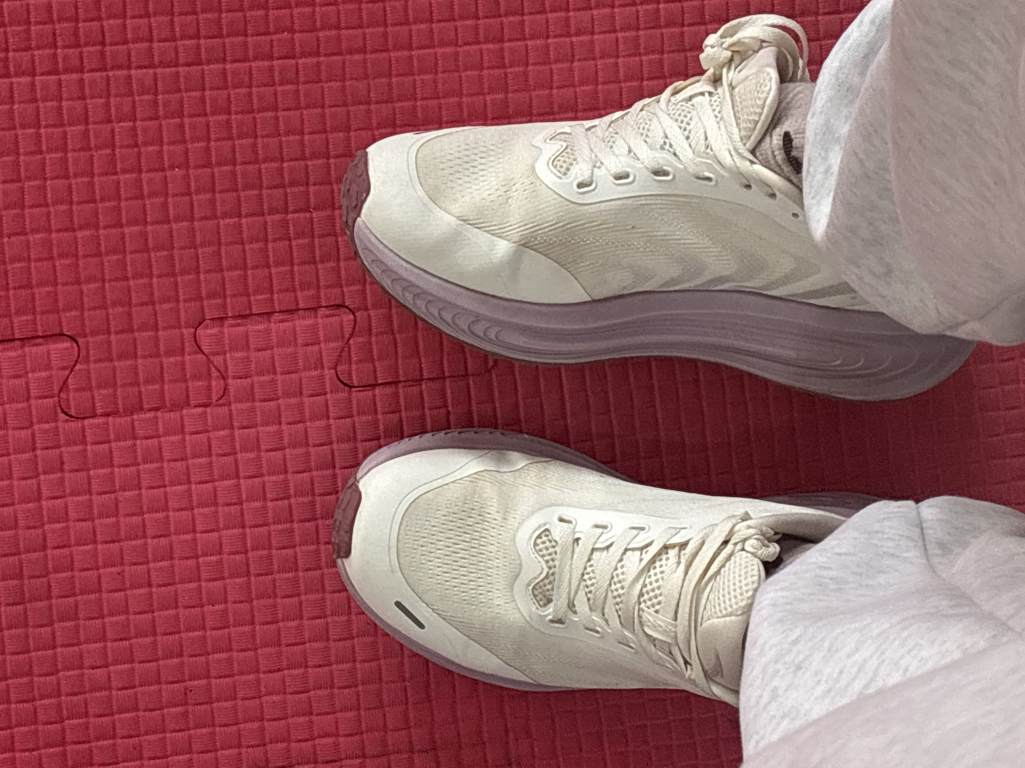 The scene is situated in Tayshet, Russia, within an indoor environment. The image presents a downward-facing perspective of an individual's lower legs and feet resting on a red textured foam puzzle mat. The subject is wearing light-colored, possibly off-white or light grey, athletic trousers that obscure the ankles and partially cover the upper sections of the shoes. Two athletic shoes, primarily off-white or very light beige in color, are visible. These shoes feature pale lavender or light purple soles/midsoles, with a small reddish-brown segment on the toe and heel areas of the outsole. The uppers are constructed from a mesh-like material, and the laces are tied. The shoes exhibit some signs of minor wear. The underlying surface is a bright red foam mat composed of interlocking puzzle pieces, characteristic of a gym floor or activity area. The mat's surface shows a fine, square grid pattern. Two distinct mat pieces are visible, with their wavy interlocking seam running from the lower right to the upper central portion of the frame. No other objects or individuals are discernible in the immediate vicinity.