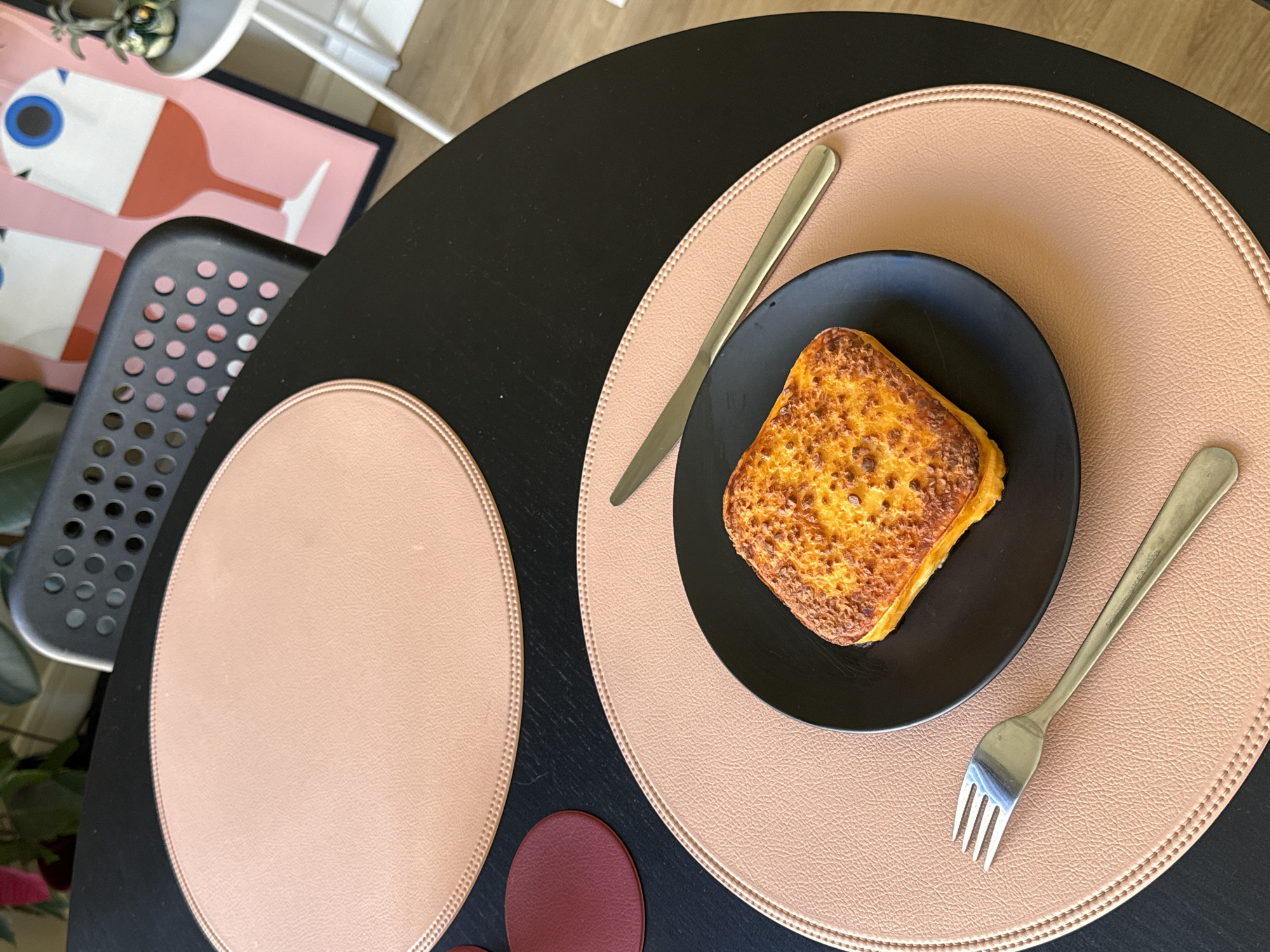 The image shows a top-down view of a meal setting, likely indoors. A black, round table is set with two light-pink circular placemats and a couple of burgundy coasters. On one of the placemats is a black plate with a slice of what appears to be French toast or bread pudding, and a fork and knife. A chair with a perforated seat is partially visible on the left side of the frame. In the background is a framed print showing an abstract design. This scene appears to be set within a modern residential setting. The location in Barcelona, Spain, is not verifiable from the image, and there is no evidence of any people, actions, or interactions in the provided content.
