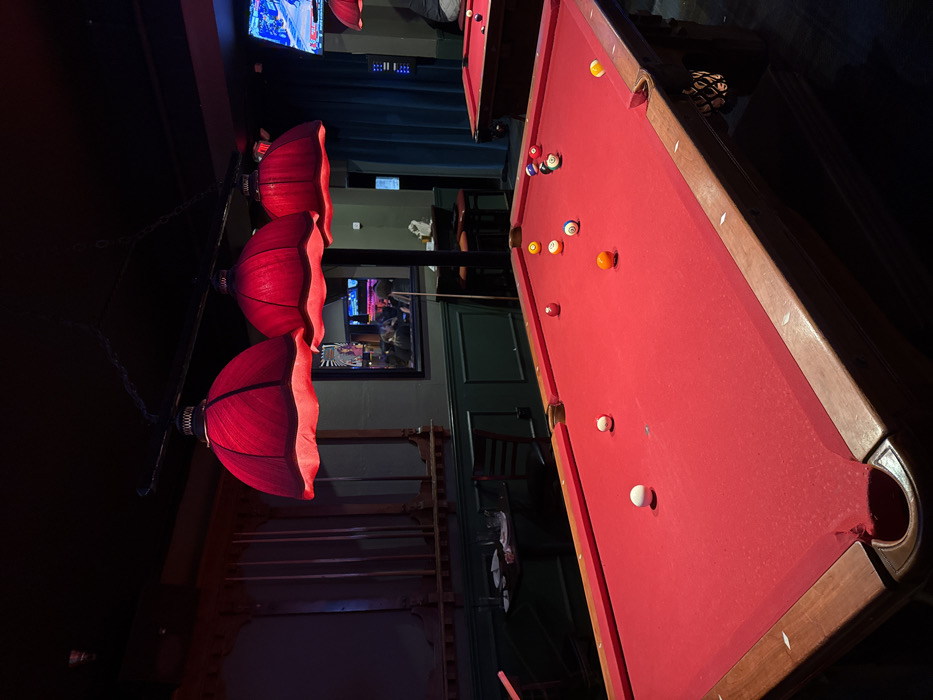 The image displays the interior of a dimly lit commercial establishment, identified as a pool hall or bar, located in Los Angeles, California, United States of America.
In the foreground, a rectangular pool table with red felt and wooden rails occupies the majority of the frame. Ten billiard balls are scattered across its surface: two white balls (one near the center-left, another near the bottom-left corner pocket), one solid red ball (upper-left), one solid yellow ball (upper-center), one solid orange ball (mid-right), one solid green ball (upper-right), one striped yellow ball (upper-right), one striped blue ball (mid-right), one striped green ball (upper-right), and one striped purple ball (upper-right). Directly above this table, three red, inverted bell-shaped fabric lampshades are suspended, illuminating the playing surface.
To the right, a second pool table with red felt is partially visible in the mid-ground, showing at least two balls (one yellow, one red) on its surface. Another red lampshade hangs above this table. The walls are painted a dark green. To the left, an ornate wooden paneling section features a rack holding multiple pool cues. A small, square dark table with two glasses and a dark chair is positioned against the left wall.
Further back, a large mirror on the wall reflects indistinct figures of several individuals within the establishment and a pool cue leaning against the wall. To the far right, a flat-screen television is mounted, displaying content with prominent blue and white colors, indicative of a sports broadcast. Dark teal curtains hang adjacent to the television. The ceiling is dark, and the overall ambient lighting is low. No explicit actions or interactions are discernible from the partially visible individuals.