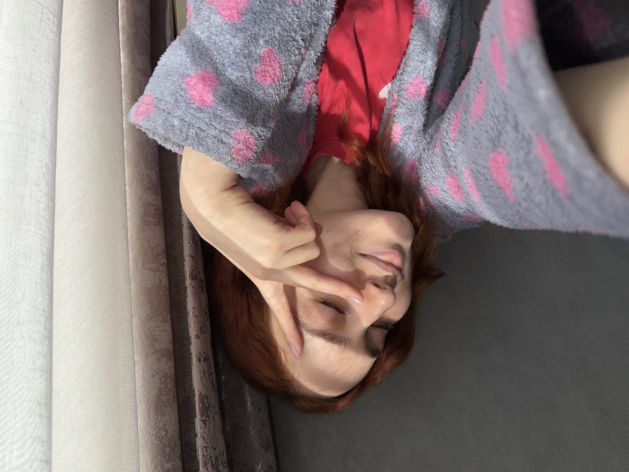 A woman is lying on a dark gray surface in front of a window with light and darker gray curtains. She is wearing a red shirt and a gray robe with pink polka dots. Her auburn hair is visible around her head. She is holding her hand up to her face. The location cannot be determined from the given image.