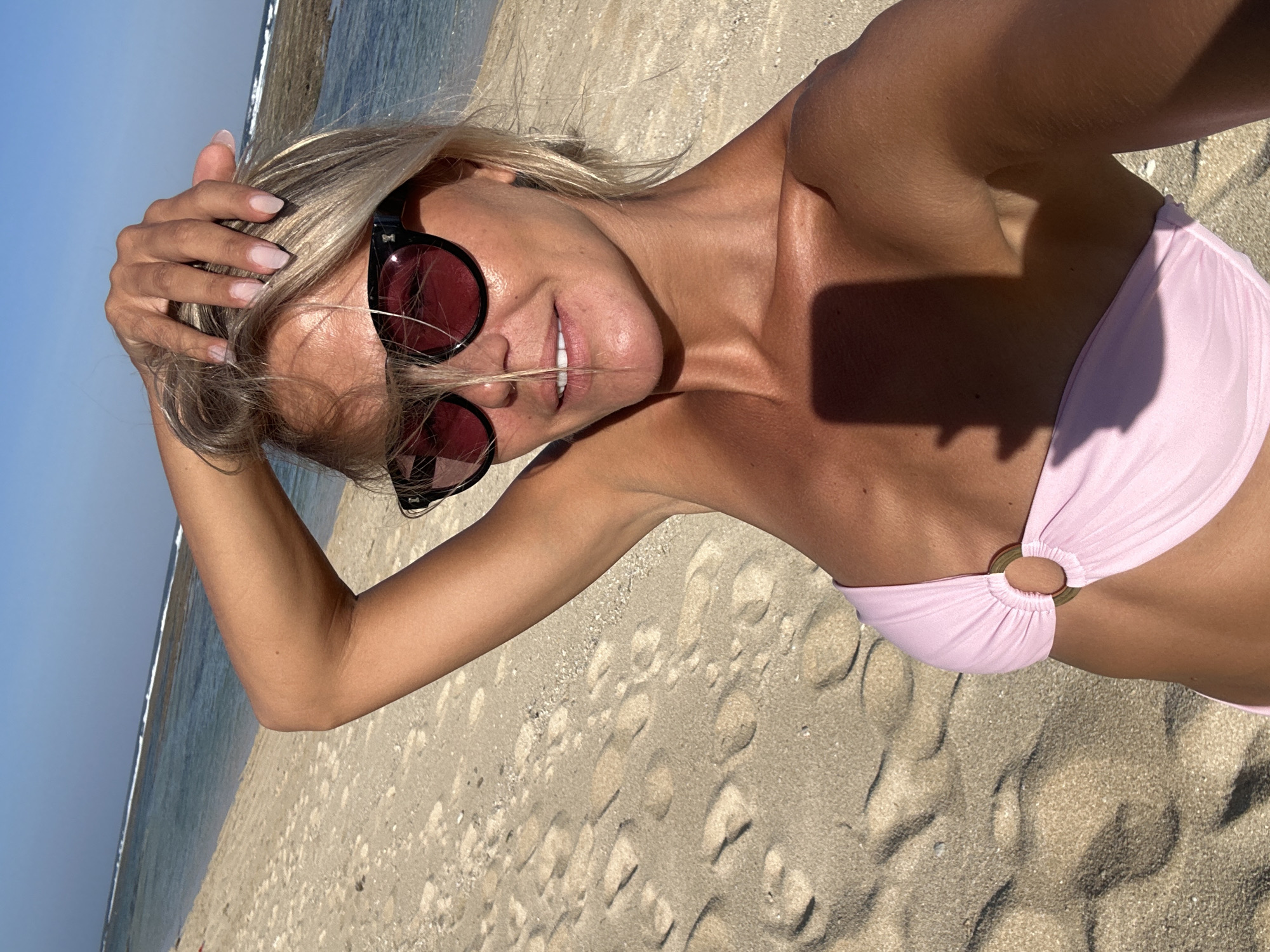 A woman is taking a selfie on a beach in Kuta Selatan, Indonesia. She is wearing a light pink bikini and sunglasses with red lenses. Her hand is raised to her head, and her blonde hair is partially covering her face. The sun is shining brightly, casting shadows on the sand and her body. The ocean is visible in the background, with waves gently lapping at the shore. The beach is sandy with footprints. The woman appears relaxed and happy, with a slight smile on her face. The time of day is likely midday, given the strong sunlight and shadows.