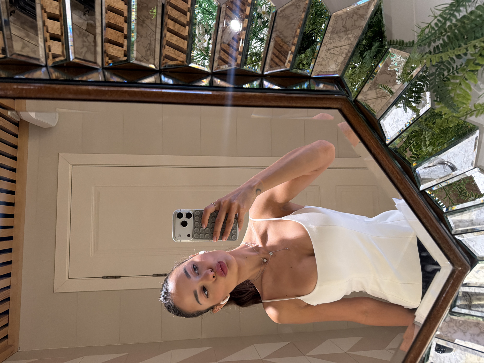 A female individual is depicted in a mirror selfie within an indoor setting. She is positioned to the left of a large, ornate mirror with a dark wooden frame and numerous beveled, angled reflective segments. She has dark hair pulled back and is wearing a white, scoop-neck top with thin straps. White wireless earbuds are visible in both ears. She wears two silver necklaces: a shorter one with a square-shaped pendant and a longer, drop-style necklace featuring a single round pendant. A small crescent moon symbol tattoo is visible on her left upper arm. Her lips are puckered. She is holding a light-colored smartphone with a grey, textured case in her left hand, using it to capture her reflection.
The reflection directly behind her reveals a light-toned wall with horizontal paneling or shiplap. A white interior door with a single visible hinge is prominent on the left side of the reflection. Above the wall and door, a wooden slatted ceiling is visible. To the right of the door, a white wall-mounted light fixture is present.
The numerous angled segments on the right side of the mirror frame reflect parts of the room behind the camera, primarily displaying multiple reflections of lush green artificial foliage, including ferns and small white flowers, suggesting decorative plants. These segments also show fragmented views of the wooden slatted ceiling.
The specific location within Moscow, Russia, is not identifiable from the visual information presented in the image.