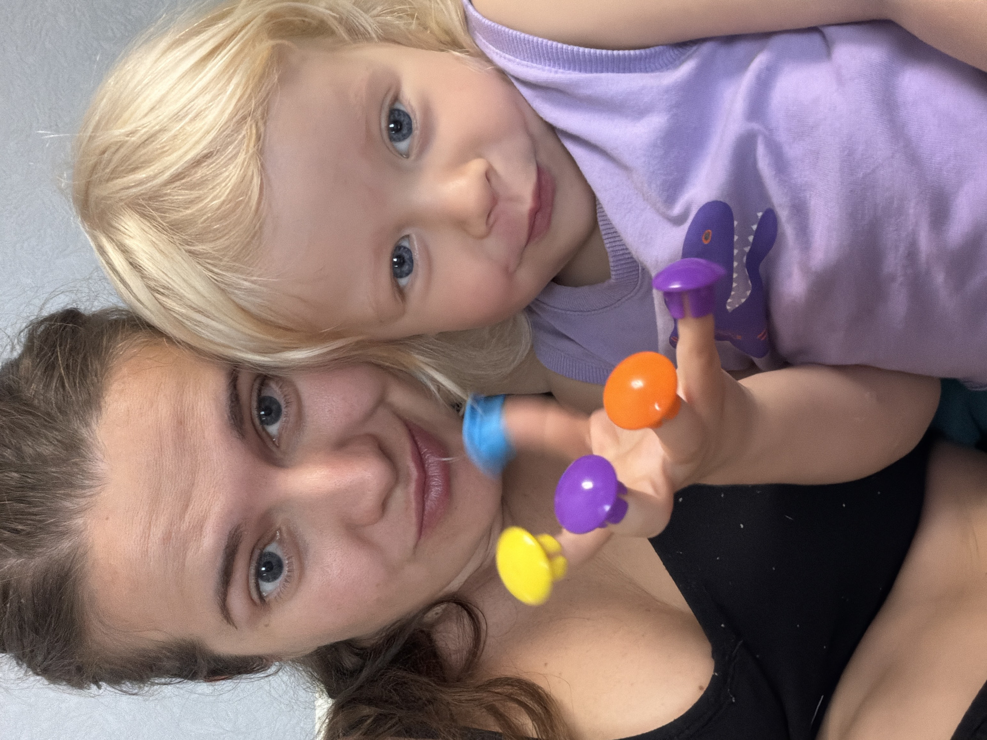 A mother and her young daughter are positioned close together, appearing in a selfie-style photograph. The mother has light skin, brown hair, and blue eyes. The daughter has fair skin, blonde hair, and blue eyes. The daughter is wearing a light purple shirt with a purple dinosaur emblem. The mother is wearing a black top. The daughter's hand is visible, adorned with colorful finger puppets in blue, orange, purple, and yellow. The background is a plain light gray wall. There is no evidence to support the location being Sochi, Russia.