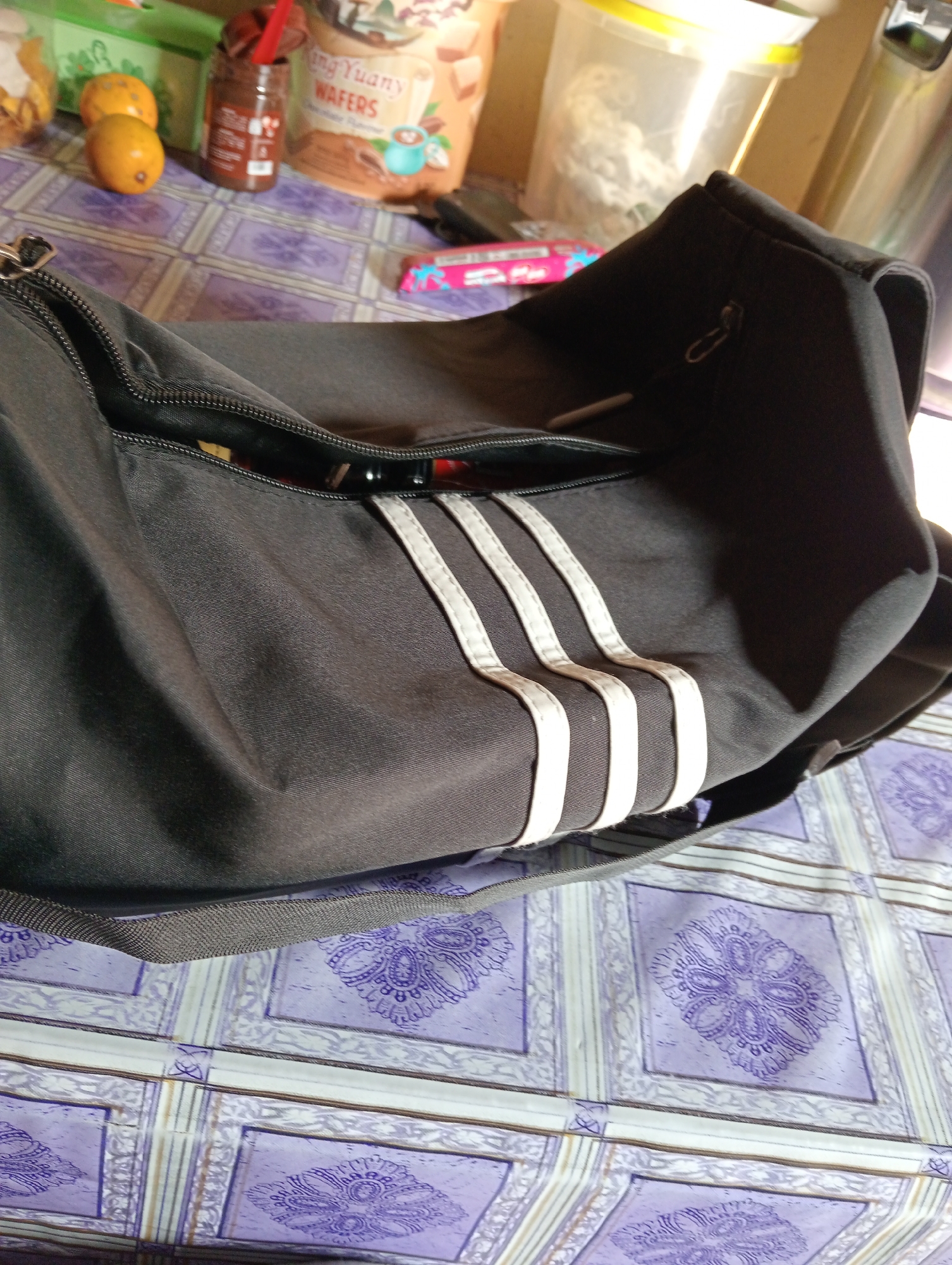 The image shows a close-up of a black duffel bag with white stripes, resting on a patterned surface. The bag is partially unzipped, revealing some items inside. In the background, on a table, are two oranges, a jar of chocolate spread, and a package of wafers. The patterned surface appears to be a tablecloth. The setting seems to be a domestic environment, possibly a kitchen or dining area. The image is taken from a high angle, looking down at the bag. There are no people visible in the image. The lighting is natural, and the overall impression is one of everyday life.