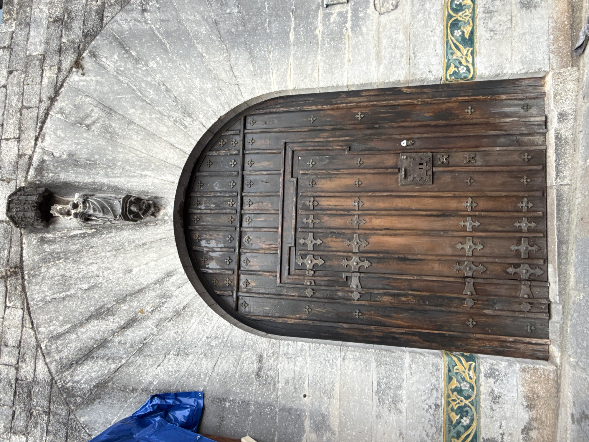 This is a low-angle, eye-level, medium shot of a large, dark wooden arched door set into a stone facade. The door is composed of vertical planks and is heavily ornamented with dark metal decorative elements, including crosses and what appear to be hinges. A heavy, ornate metal lock is visible on the right-hand door panel. The stone archway above the door is constructed from rough-hewn, light-colored stones radiating outwards from the top. Mounted centrally above the arch, on the stone facade, is a dark stone carving of a robed figure, possibly a saint, holding an object in its arms, with a smaller figure at its feet. The stone wall to the left of the arch has a section covered by a bright blue tarp. Below the arch and to either side of the door, a decorative tile band with a repeating green and yellow floral pattern is visible. The foreground shows a rough stone step. The image appears to be of the entrance to a building with historical or religious significance, possibly located in Girona, Spain, given the architectural style and decorative elements.