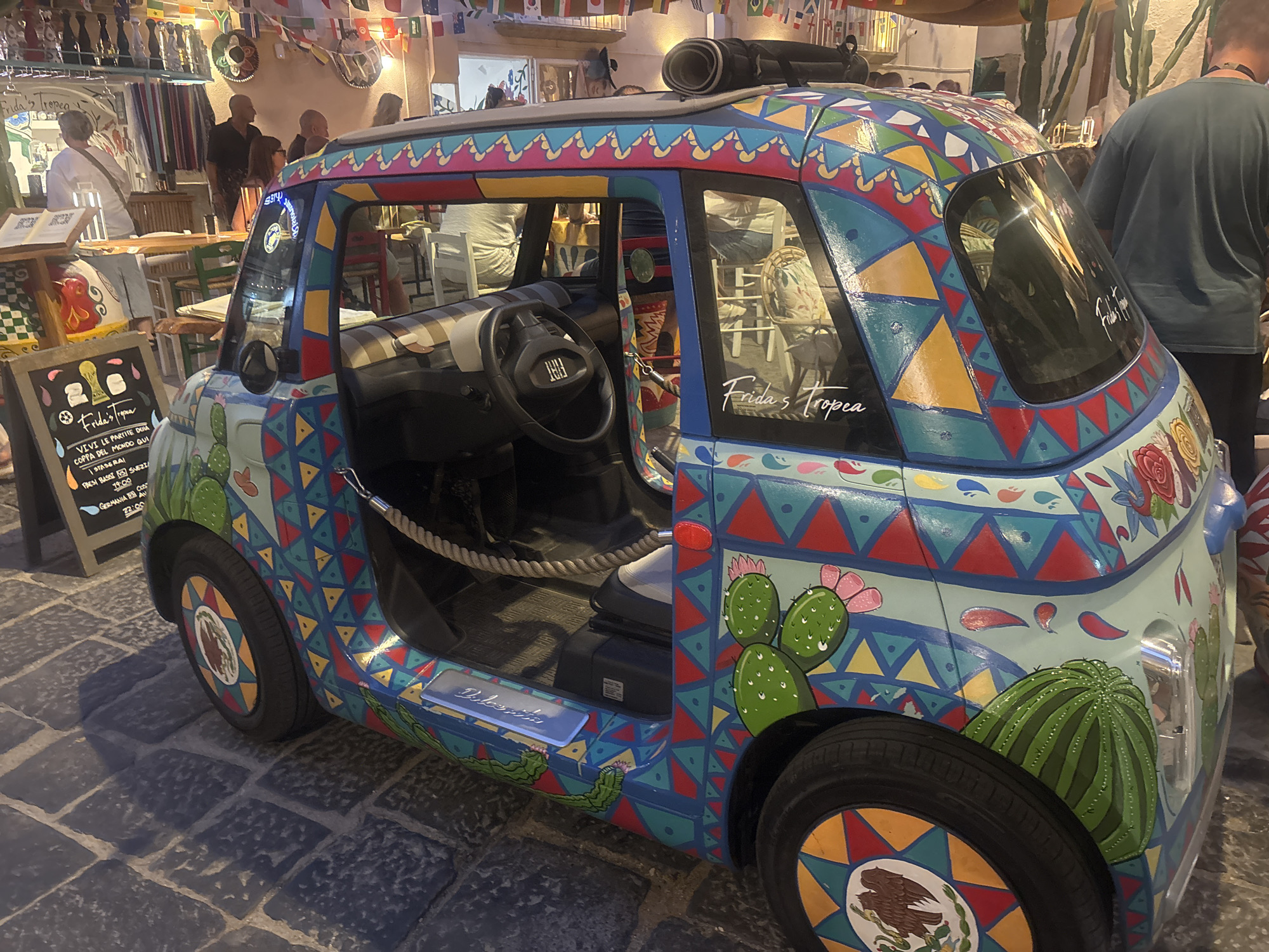 In Tropea, Italy, a custom-painted Fiat 500, named "Frida's Tropea," is parked on a cobblestone street, adorned with vibrant Mexican-inspired folk art, including cacti and geometric patterns. The car's interior, visible through the open driver's side door, reveals a steering wheel and seating. To the left of the car, a chalkboard menu displays "Frida's Tropea" in colorful lettering, along with various food and drink items and times. In the background, people are seen in an establishment with tables and chairs, suggesting a restaurant or cafe setting. Flags hang from the ceiling, adding to the festive atmosphere. A person in a dark t-shirt stands to the right of the car.