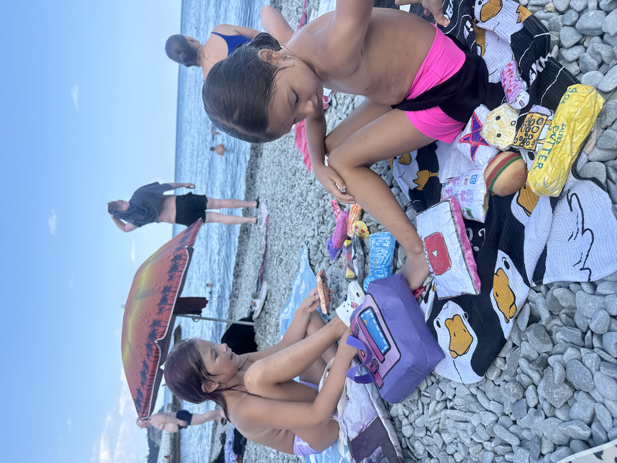 Two young girls are sitting on a pebble beach in Partenit, Ukraine, near the Black Sea. The girl on the left, with purple hair, wears a light purple swimsuit. She is looking down at a purple bag and various paper toys spread out on a black and white towel with duck imagery. The girl on the right, with dark hair, wears a pink and black swimsuit. She is looking down at a collection of colorful paper toys. In the background, an orange umbrella shades a part of the beach, and several other people are visible, some in the shallow water. The sky is blue with scattered clouds.