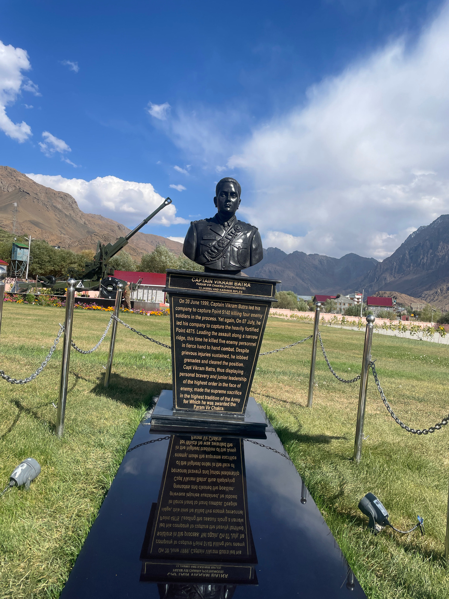 The image shows a bust of Captain Vikram Batra, a posthumous recipient of the Param Vir Chakra, placed in front of a monument with an inscription detailing his bravery during the Kargil War. The monument is situated in a grassy area with a mountainous backdrop under a clear blue sky with some clouds. A military cannon is visible to the left of the monument, and a chain-link fence surrounds the memorial. The overall scene appears to be a war memorial or museum exhibit in Bhimbat, India.