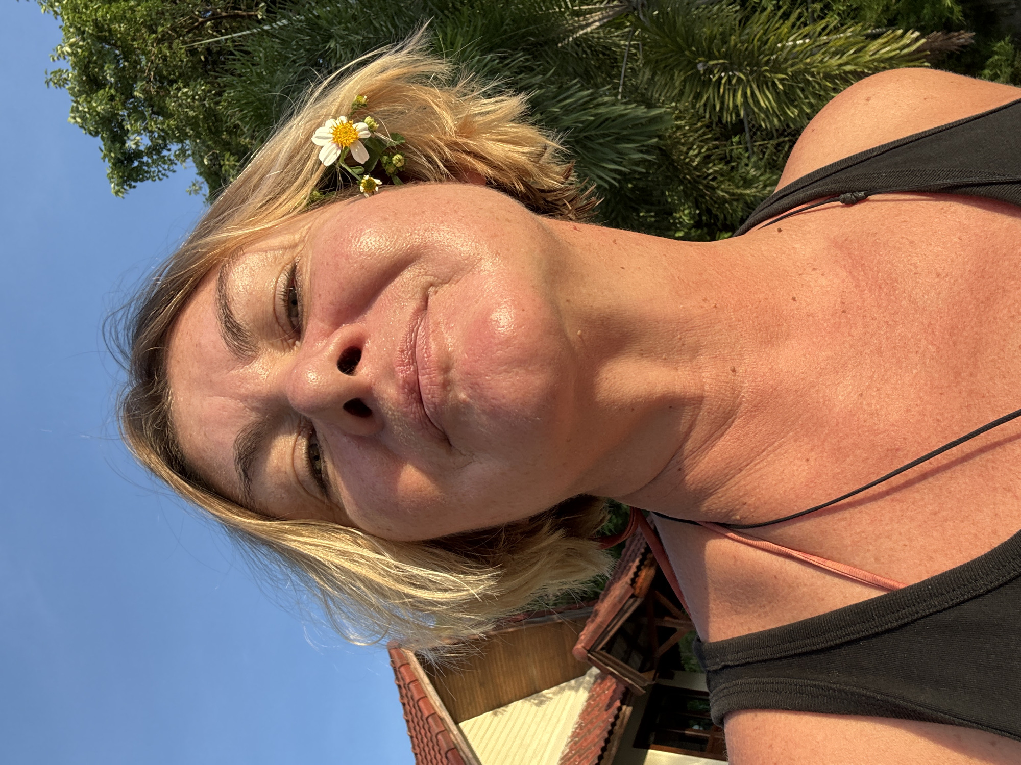 A close-up selfie of a blonde woman adorned with a small white flower in her hair. She is wearing a black tank top, and her skin appears sun-kissed. The background shows lush green foliage, suggesting a tropical or outdoor setting. A glimpse of a building with a terracotta-tiled roof can be seen beneath the greenery. The sky is a clear, bright blue, indicating daytime with good weather. The woman's expression is relaxed and content.