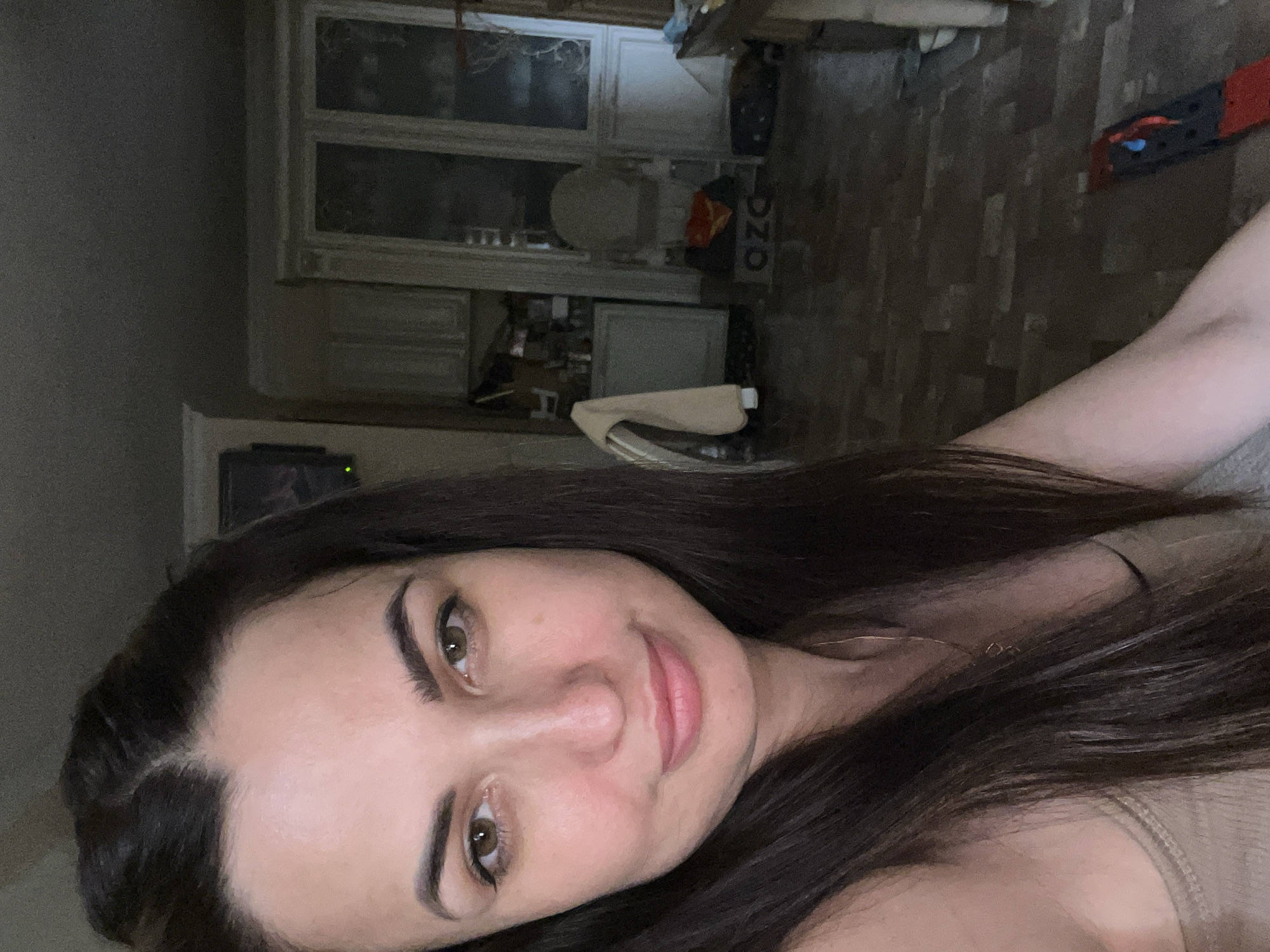 A woman with long, dark hair and green eyes is taking a selfie in what appears to be a home environment. She is wearing a beige tank top and a gold necklace. Her hair is pulled back from her face. In the background, there are kitchen cabinets, a glass-paned door, and a chair. The flooring is a patterned tile or laminate. The lighting is dim, creating a slightly shadowed atmosphere.