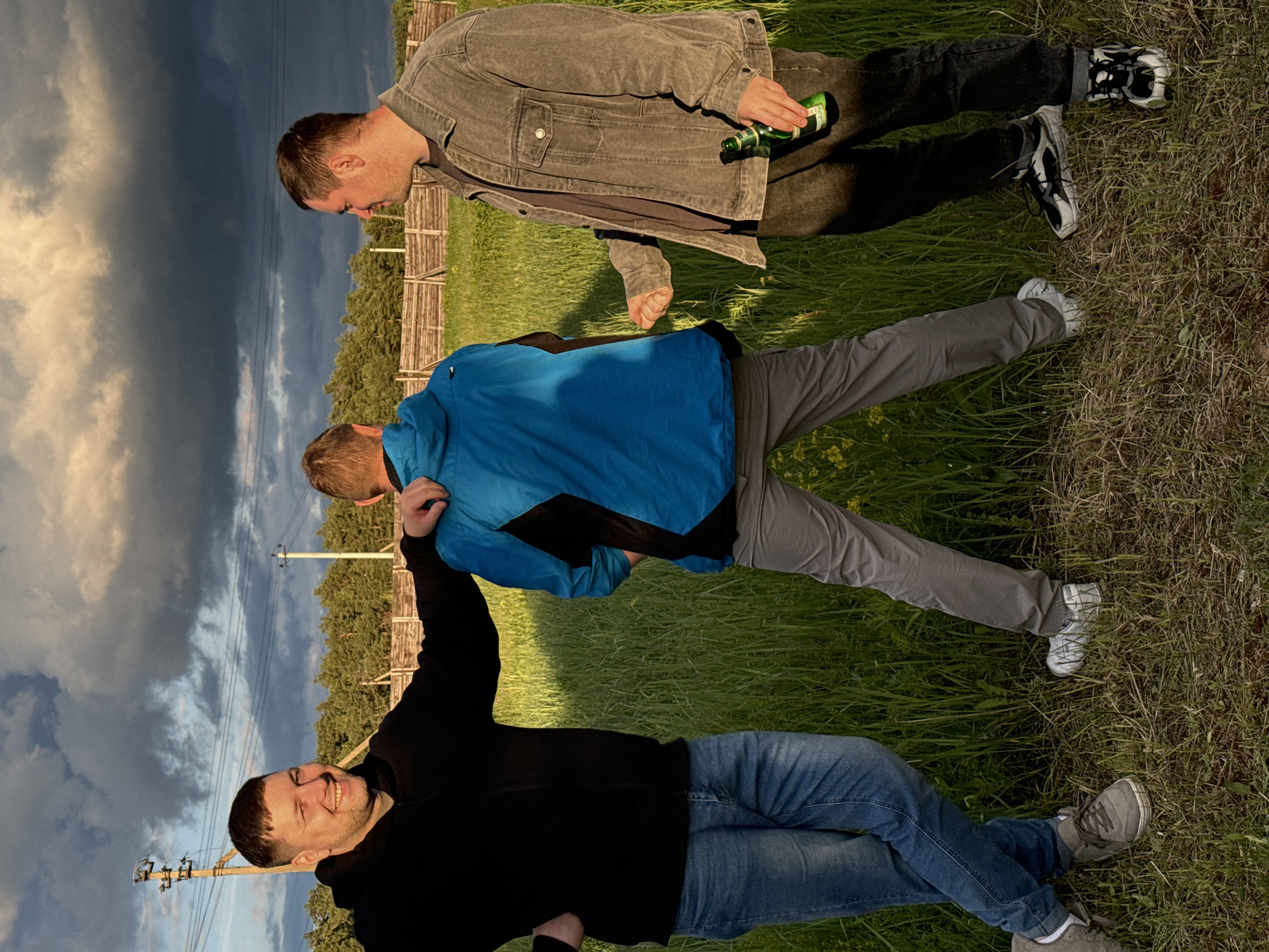 The image shows three men in a field in Vysokinichi, Russia. The man at the top wears a gray jacket, black pants, and holds a green bottle. The middle man wears a blue jacket and gray pants. The bottom man wears a black jacket and blue jeans and has his arm raised. The background includes a cloudy sky, trees, and power lines. A wooden fence is visible between the top and middle men. The men are positioned vertically in the frame and are lying down.