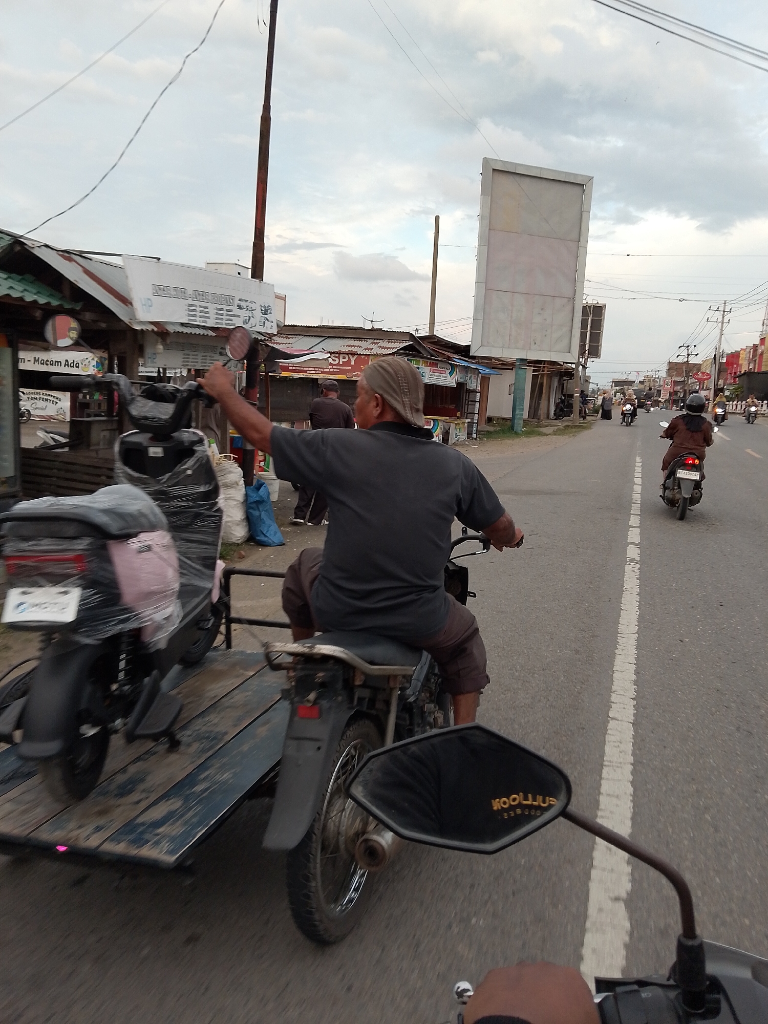 An overcast day in Meunasahdayah, Indonesia. A person wearing a grey t-shirt and brown shorts, with a light-colored head covering, is riding a motorcycle while pulling a trailer carrying two other motorcycles. The trailer is wooden and weathered, with the motorcycles wrapped in clear plastic. The person in the foreground is looking to their right, with their right arm extended. Other motorcycles and people are visible on the road in the background, with buildings lining the street. A large, empty billboard stands on the right side of the road. The scene appears to be a street in a commercial or residential area.