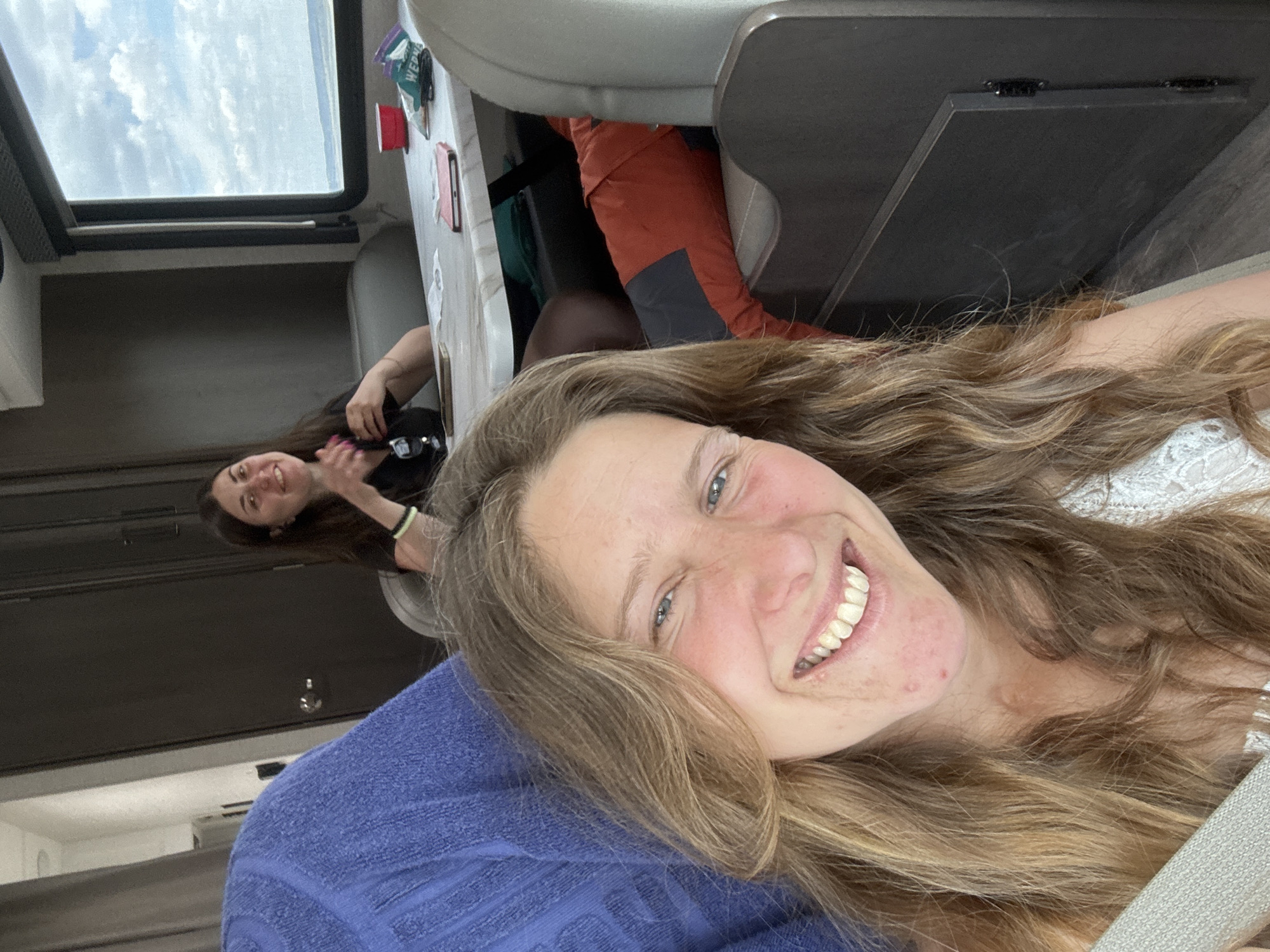 The image shows two women inside what appears to be a recreational vehicle (RV). The woman in the foreground is smiling broadly at the camera, lying against a blue material. Her hair is long and wavy, and she is wearing a light-colored top. Behind her, another woman with dark hair is sitting at a table near a window that shows a cloudy sky. The interior of the RV is visible, with features like cabinets and seating. The location in the city of Unknown, Unknown is impossible to determine based on the provided visual information.