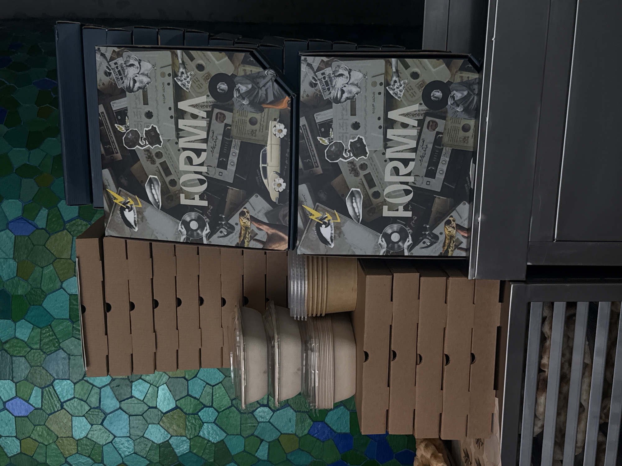 The image shows a composition of objects arranged indoors. There are two square boxes with the word "FORMA" in bold, white letters against a background of cassette tapes and other designs. These boxes are stacked atop brown cardboard boxes with small circular cutouts. Next to these are stacked disposable cups and white lids. The objects rest on a surface with a textured, mosaic-like pattern in varying shades of green and blue. A metal shelf or structure frames the objects on the right side of the image. The photo appears to be taken indoors, possibly in a commercial setting or storage area. Based on the text visible on the boxes, it may be a product related to arts or design, but the exact context is unclear. The location in the city of Dubai, United Arab Emirates is not evident from the image.