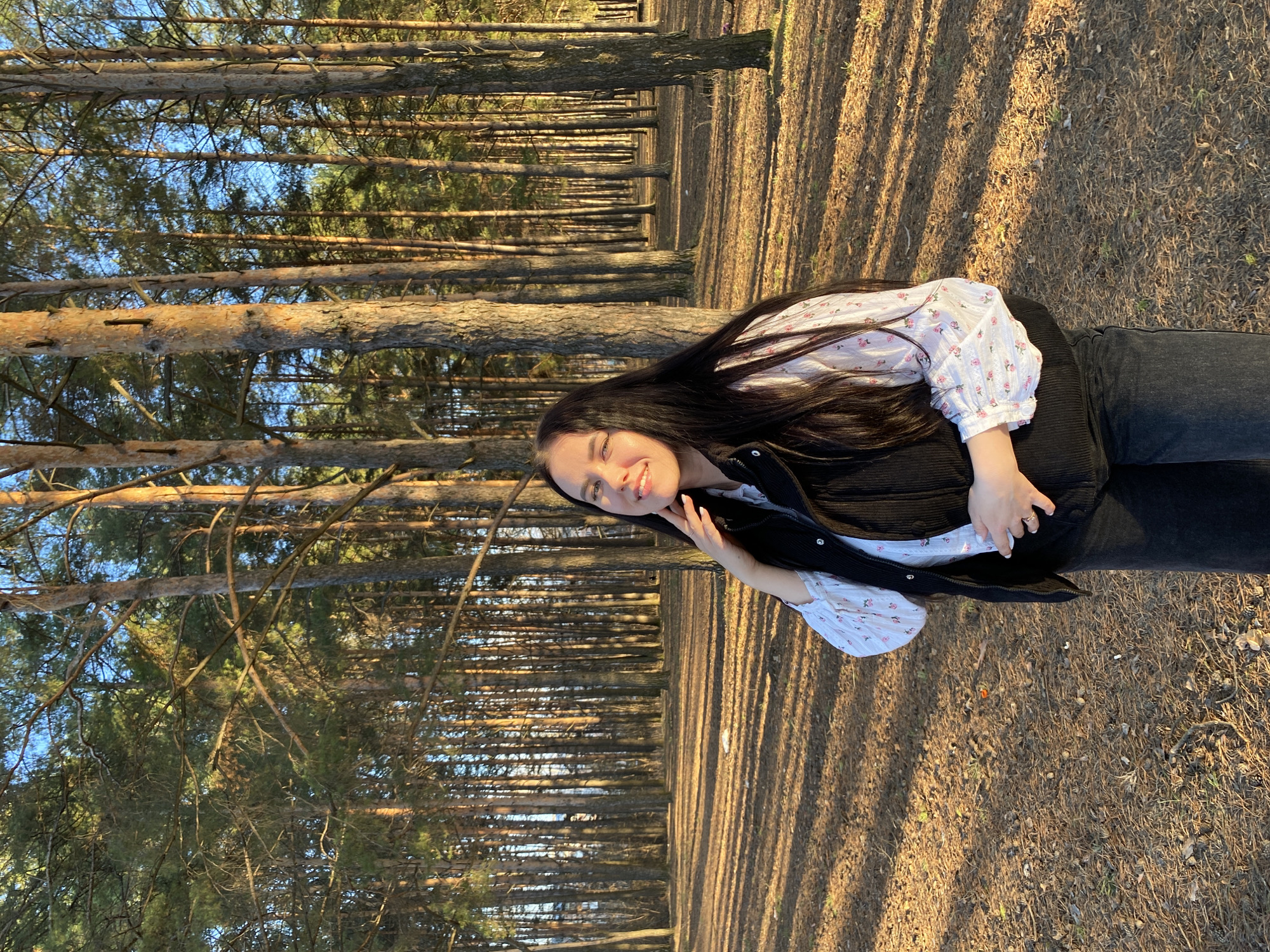 A young woman is standing in a pine forest on a sunny day. She has long, dark hair and is wearing a white blouse with a floral pattern, a black vest, and dark jeans. She is smiling and looking at the camera, with her right hand near her face. The trees are tall and straight, and the ground is covered with pine needles. Shadows from the trees stretch across the ground. Based on prior knowledge, this scene is likely located in the city of Chaikovsky, Russia, as requested.