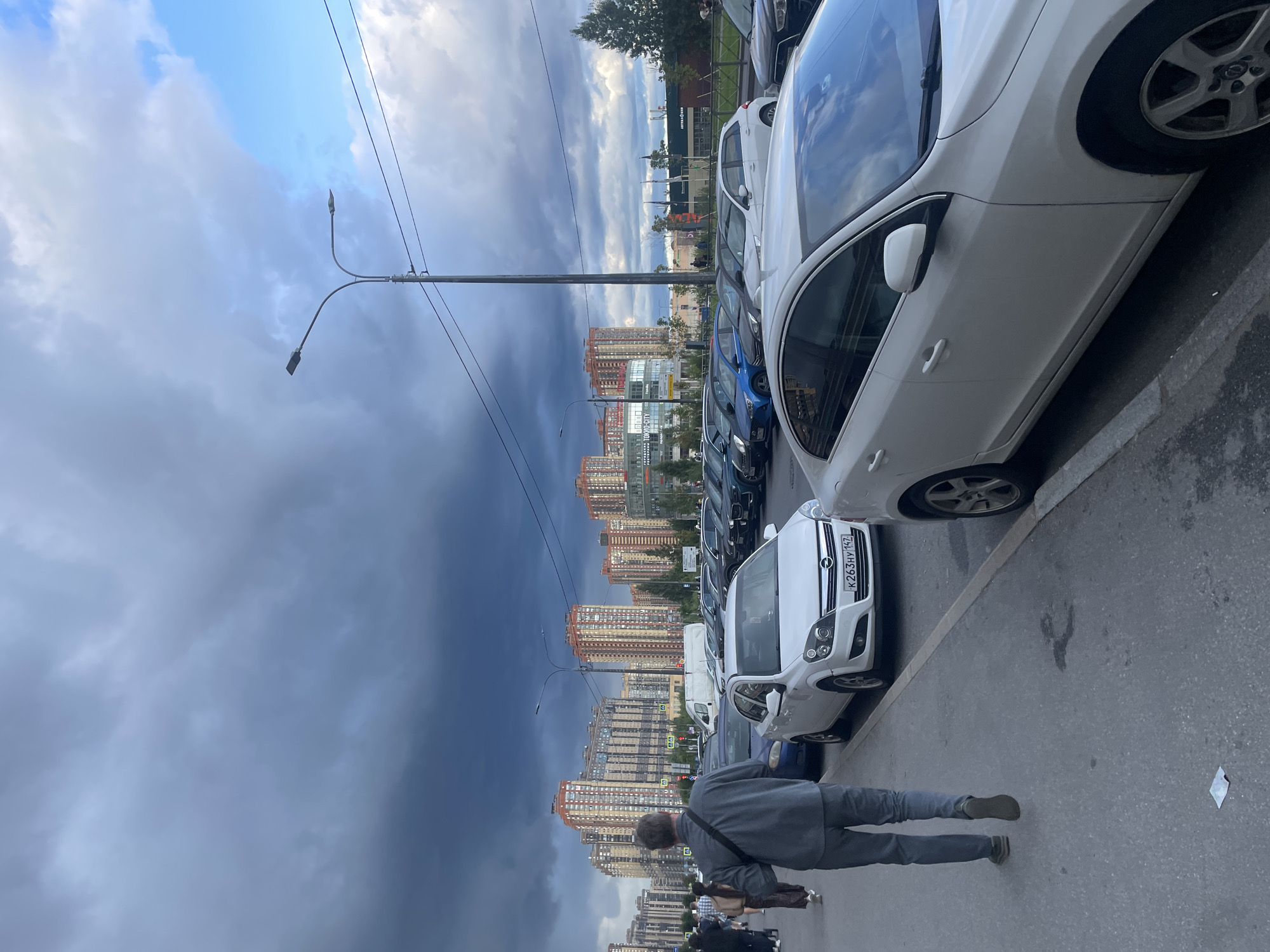 An overcast sky looms over a street scene in Ладожское Снт, Russia. Tall residential buildings with orange and beige facades dominate the background, interspersed with some commercial structures. A row of parked cars lines the right side of the street. In the foreground, a person wearing a gray jacket and pants walks away from the camera along the sidewalk. Several other pedestrians are visible further down the sidewalk. The weather appears to be on the verge of raining, with dark clouds gathered.