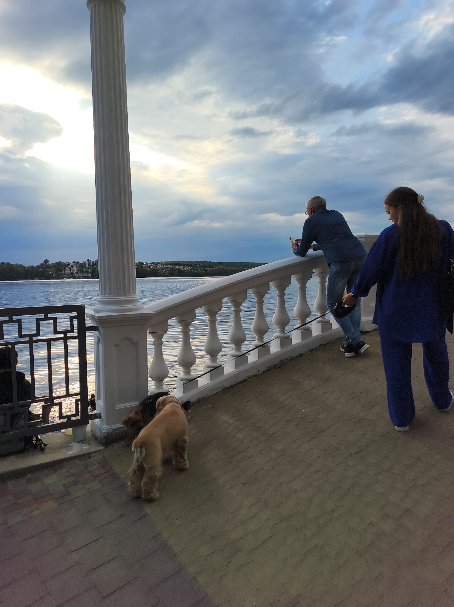 The image depicts a scenic view of Ternopil Lake in Ternopil, Ukraine, with a prominent white classical column and a white balustrade lining the waterfront promenade. The sky is overcast with dramatic clouds, and the sun is partially visible, casting reflections on the water.
In the foreground, two dogs, one light brown and one dark brown, are standing on the paved walkway. To the left, a person is seated near a black metal fence overlooking the lake, with a backpack beside them.
Further along the promenade, a man in a blue shirt and jeans is leaning against the balustrade, looking at his phone. A woman in a blue outfit is walking away from the camera, holding a leash attached to one of the dogs. The promenade is paved with rectangular bricks of varying colors. Buildings and trees are visible on the distant shoreline, indicating the presence of the city. The overall impression is a peaceful, late afternoon scene by the lake.