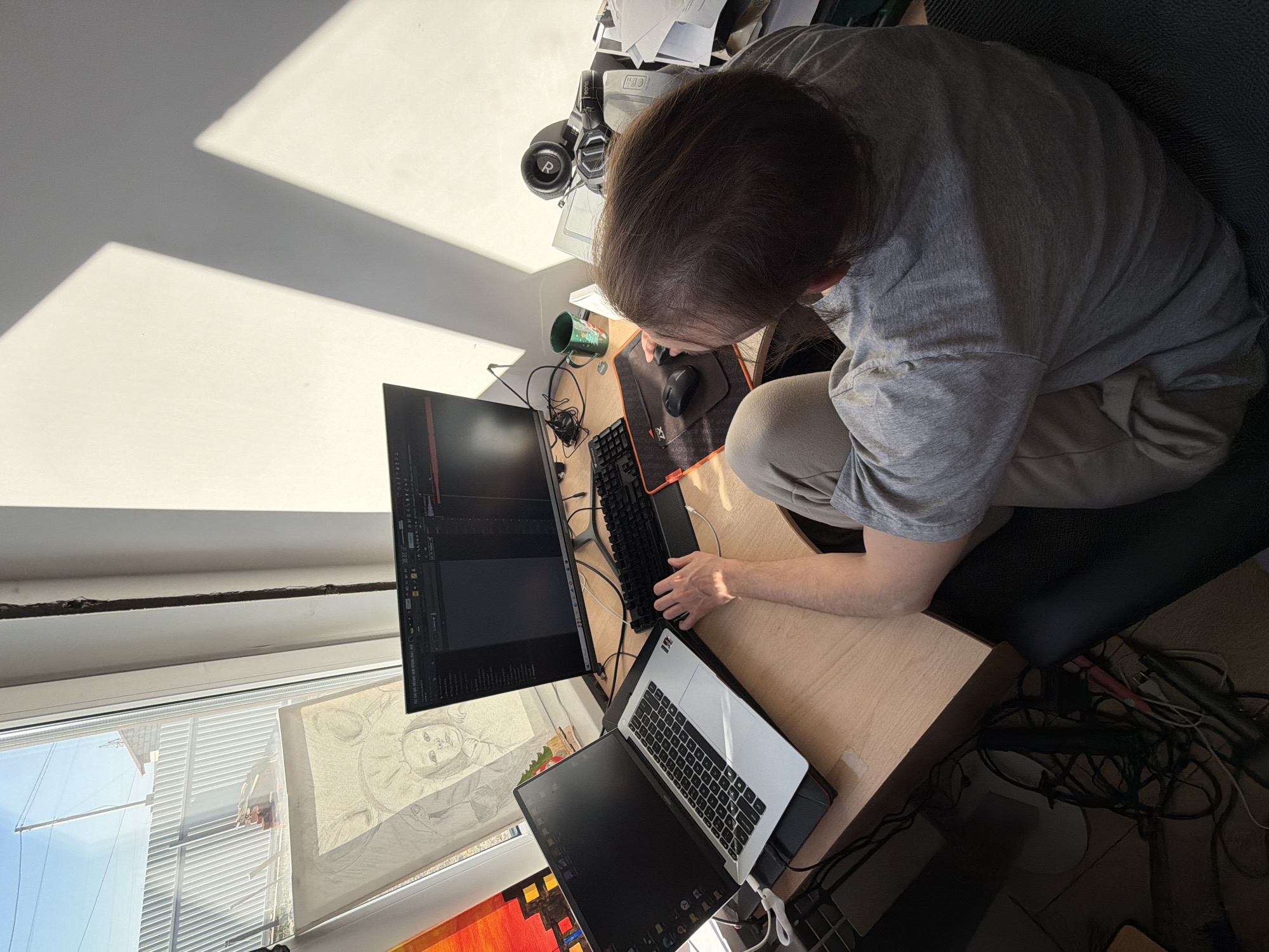 An overhead shot captures a person working at a sunlit desk in Chaikovsky, Russia. The individual, with short brown hair, wears a light grey t-shirt and beige pants, seated with their back partially to the viewer, leaning forward with focus on the computer screens.
The workspace features a wooden desk with a multi-monitor setup. On the left, a large vertical monitor displays a dark-themed interface, possibly a code editor or design software. To the right, an open laptop sits on a stand, its screen also dark. A black keyboard and a black mouse on an orange and black mousepad with "ZX" visible are positioned between the person and the monitors. A green mug with a white pattern is visible near the keyboard.
Bright sunlight streams into the room from a window on the left, casting sharp, angular shadows on the white wall, indicating a clear, sunny day. Through the window, a clear blue sky and part of a building with horizontal blinds are visible. Taped to the window frame is a light-colored drawing or print of a stylized face, possibly a baby or child. Below the window, part of a vibrant, abstract artwork with red, orange, and yellow hues is seen.
Behind the person, clutter includes black headphones with an "R" visible on one earcup, papers, and other objects. Below the desk, a dense tangle of black cables is visible. The person appears engaged in their work, utilizing the digital tools available at their desk.