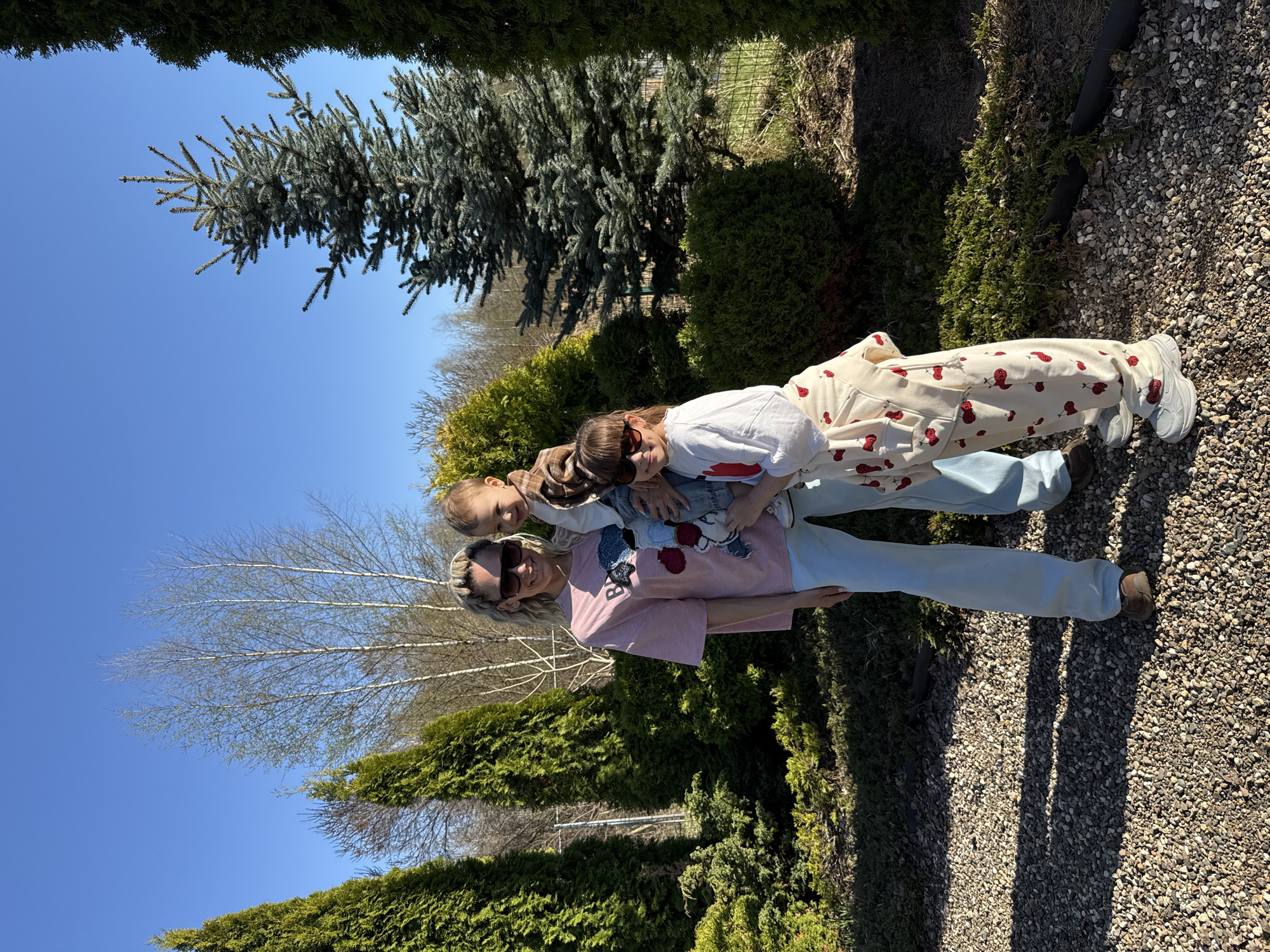 The image shows a group of three individuals standing in an outdoor setting, likely a garden. An adult woman is holding two children. The woman is wearing a light pink t-shirt, light blue trousers, and sunglasses. The children are wearing casual clothing. The environment consists of greenery, including various bushes and trees. The ground is covered in gravel. The image was taken in Rusanovo, Russia.